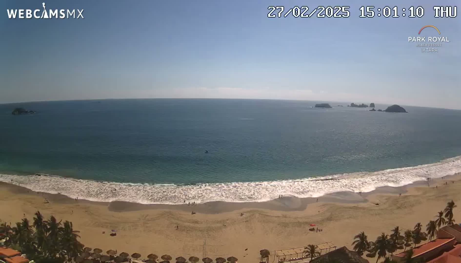 A sandy beach with gentle waves laps the shore under a clear blue sky, with small islands visible in the distance.