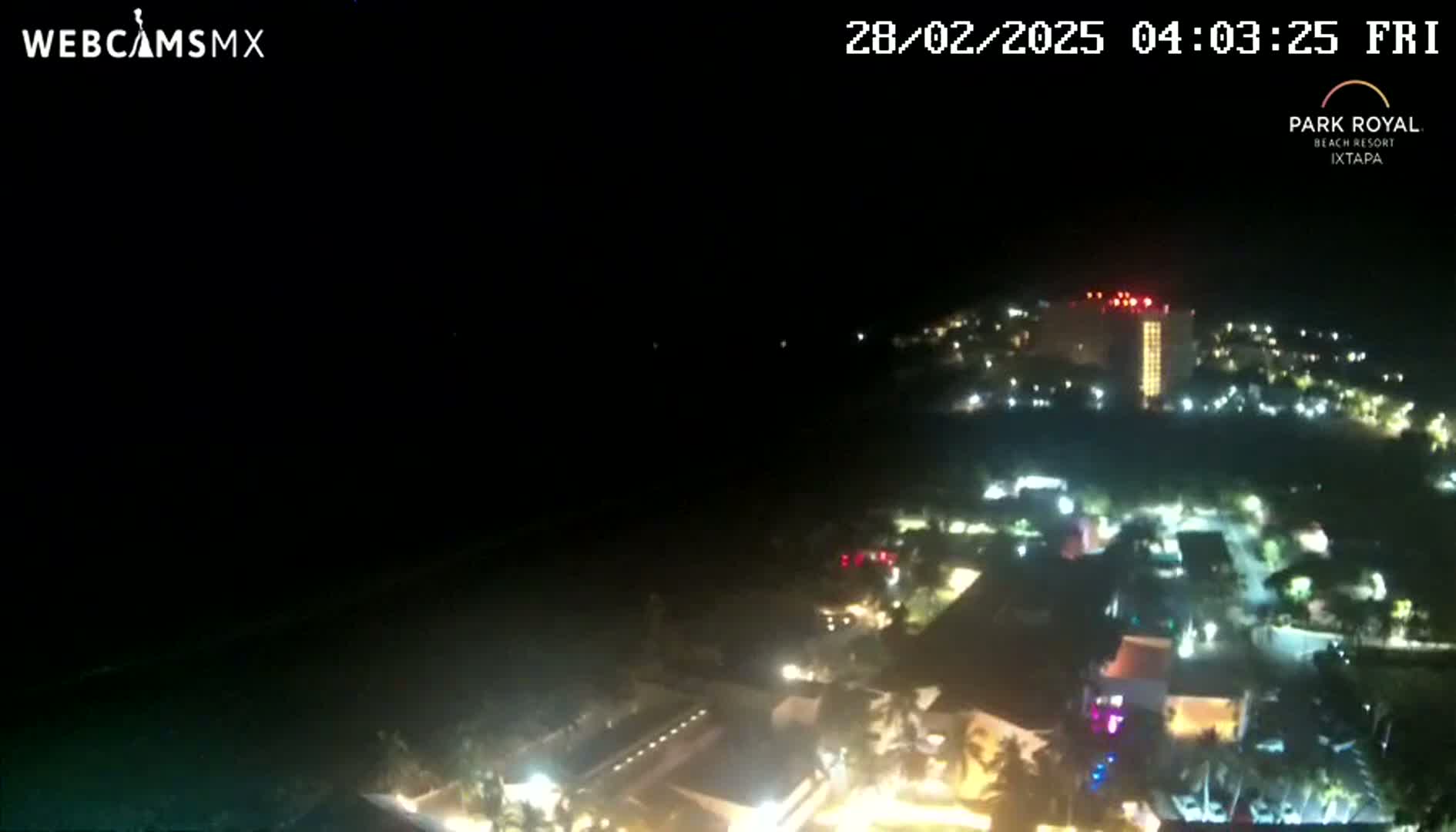 A nighttime aerial view shows a resort town illuminated by lights, with dark water visible in the foreground under a clear, dark sky.