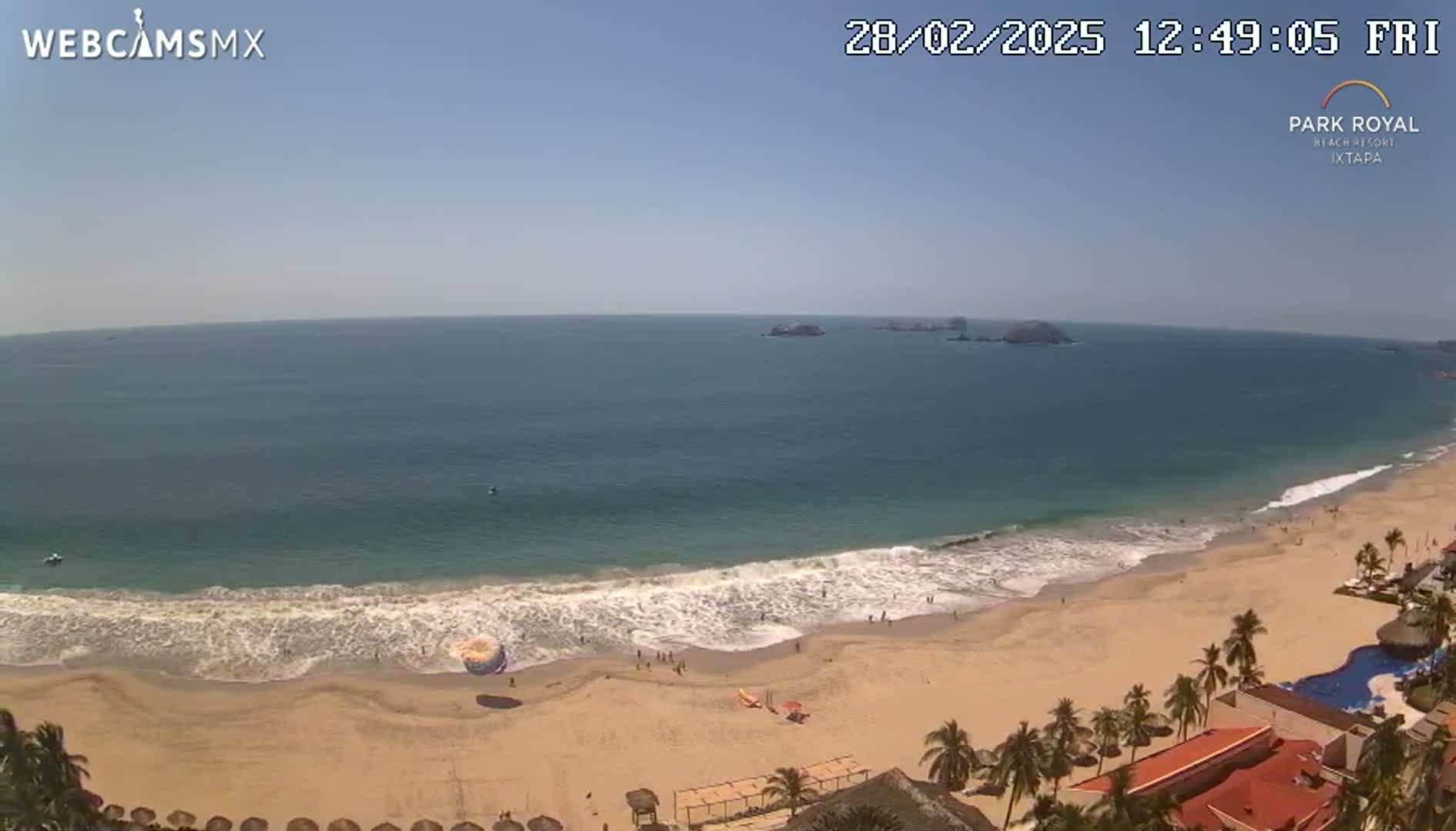 A sandy beach with gentle waves laps the shore under a clear, sunny sky, with palm trees and buildings visible in the foreground.