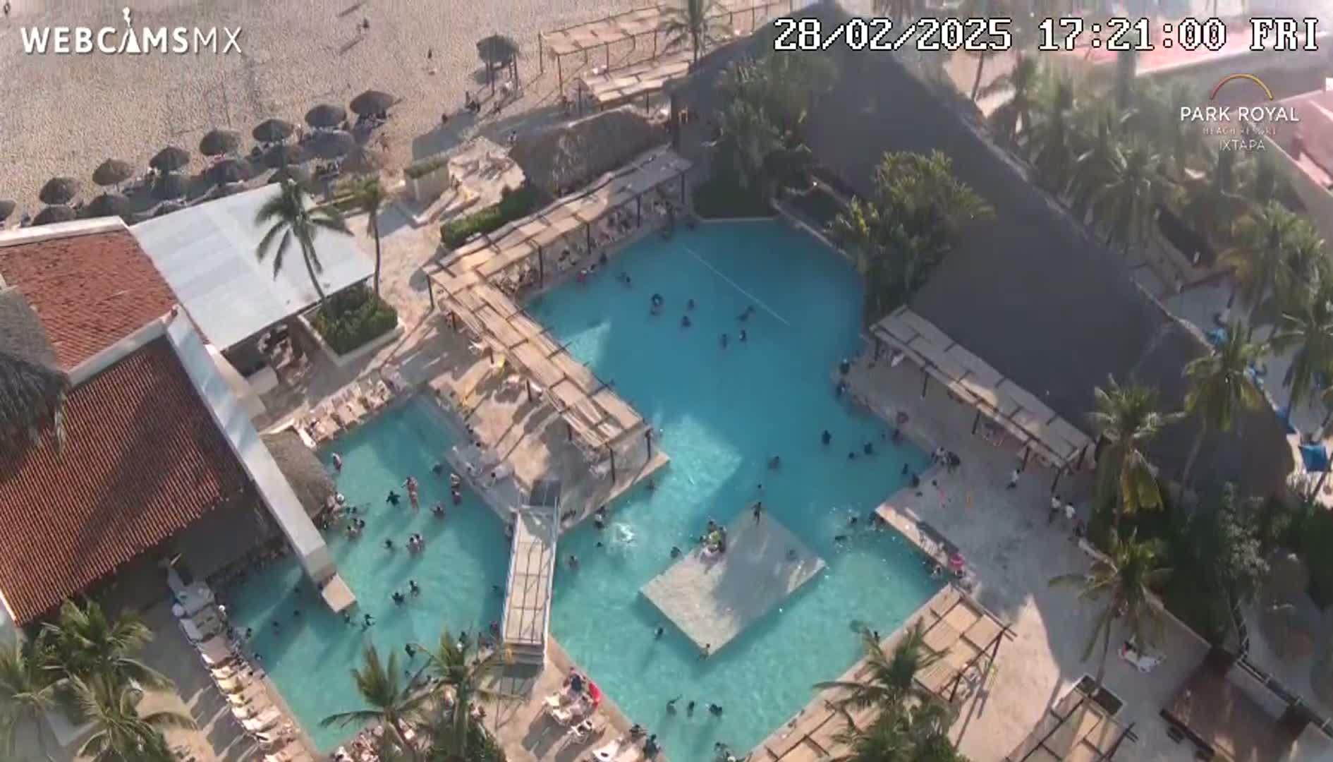 An aerial view shows a resort with multiple pools filled with people, surrounded by palm trees and a sandy beach under a sunny sky.