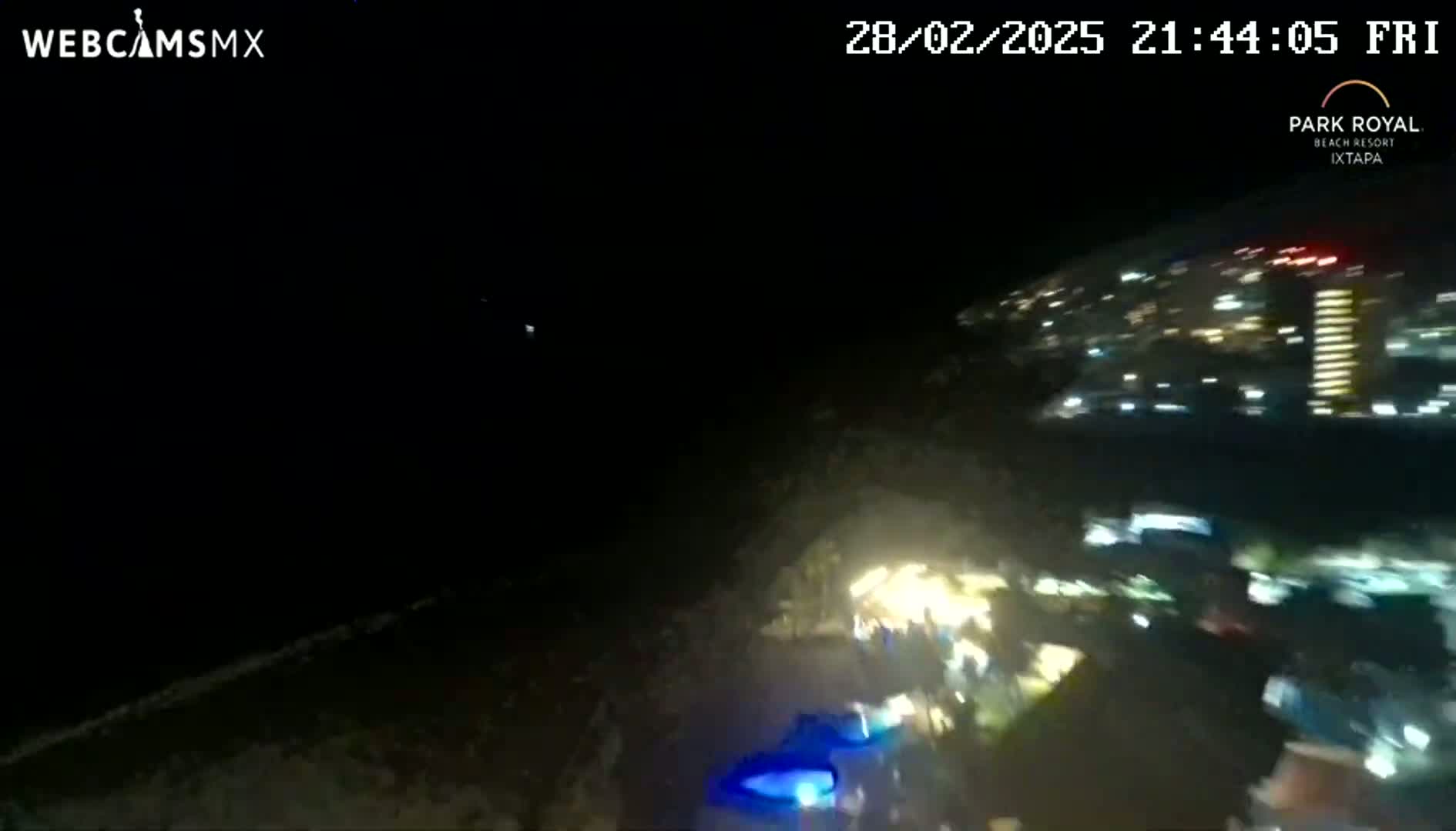 A nighttime, aerial view shows a dark coastline with a brightly lit city in the background under a clear sky.