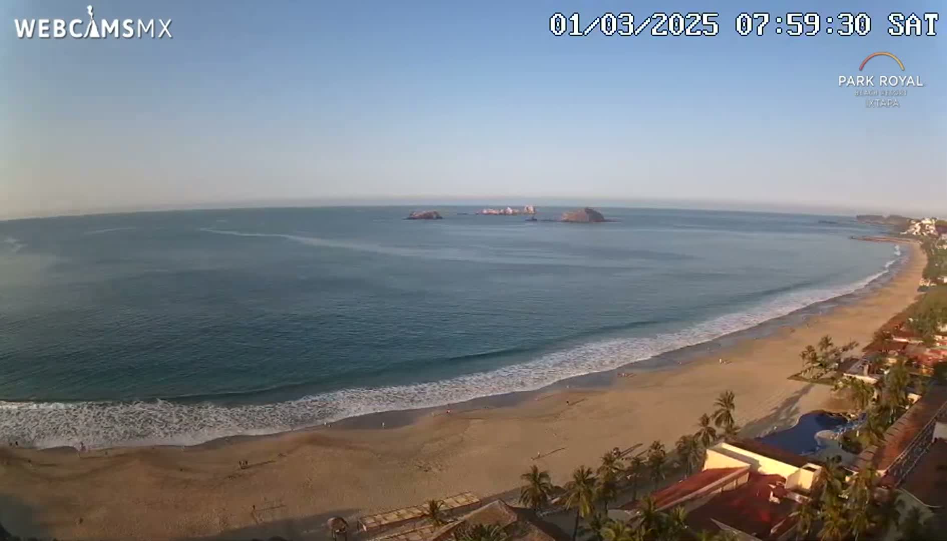 A long sandy beach with gentle waves laps the shore under a clear blue sky, with a small island visible in the distance.