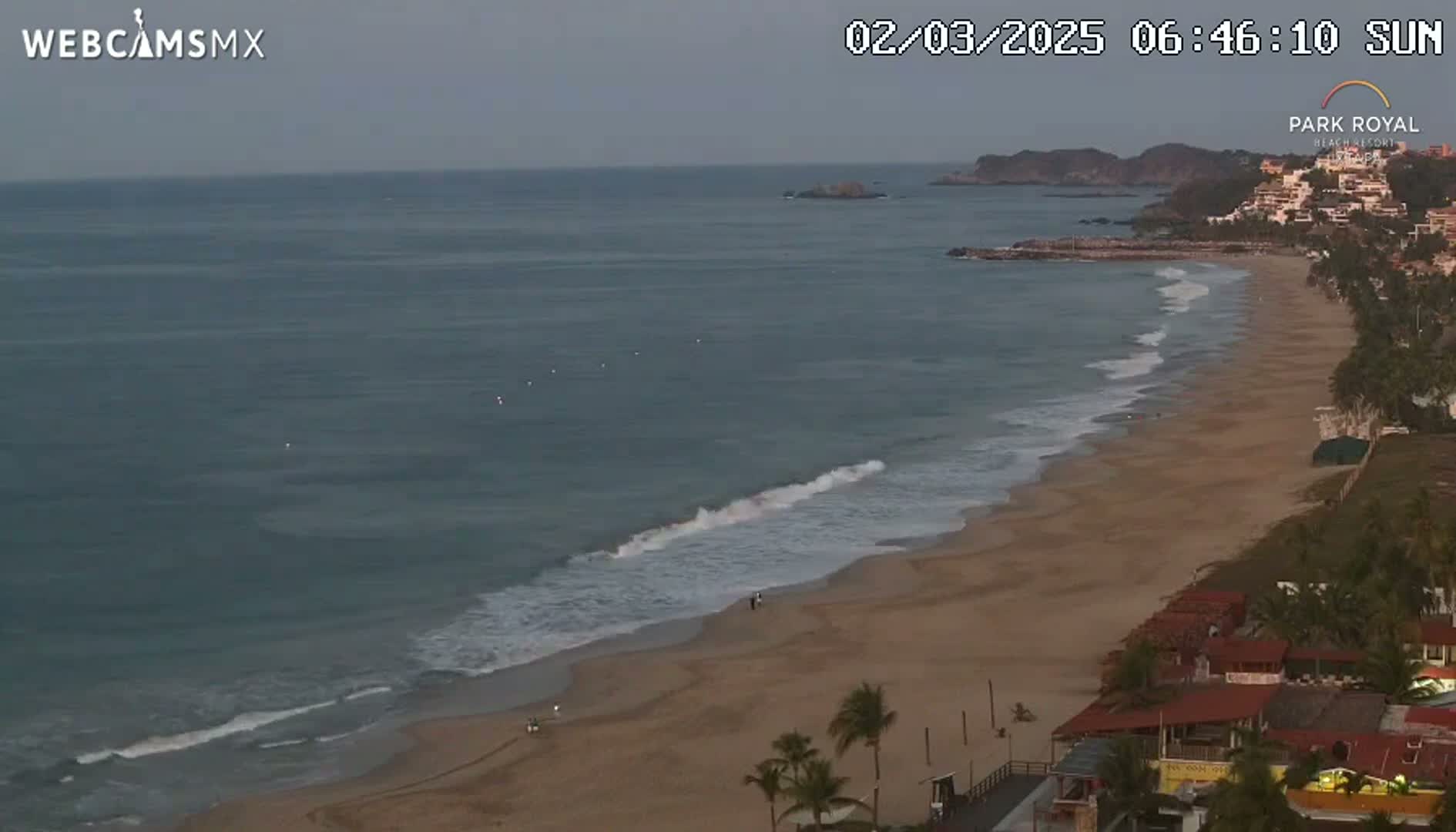 A sandy beach with gentle waves laps the shore under a mostly clear sky, with buildings and palm trees visible along the coast.