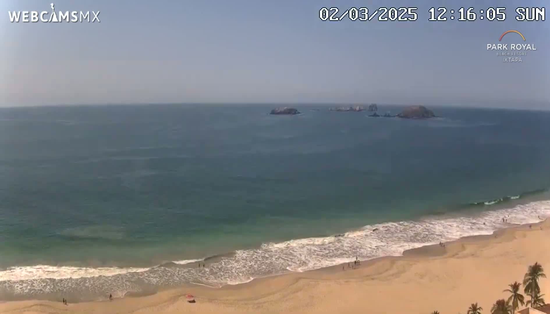 A sandy beach with gentle waves and a few people, under a clear sunny sky, overlooks a calm ocean with some small islands in the distance.