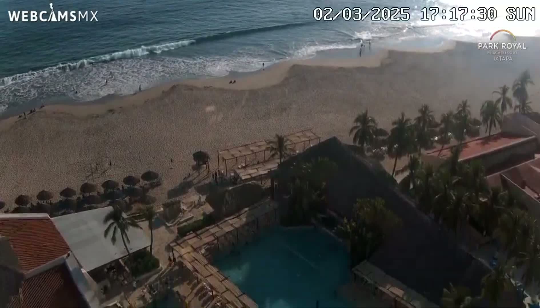 An aerial view shows a sandy beach with ocean waves, palm trees, a resort with a swimming pool, and umbrellas on the beach under a sunny sky.