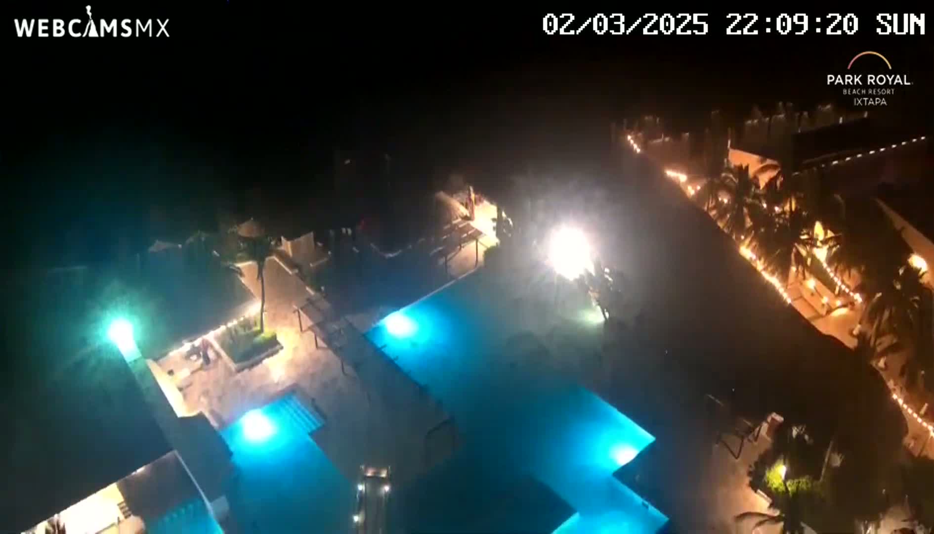 An aerial nighttime view shows two illuminated swimming pools at a resort, surrounded by palm trees and buildings with string lights.