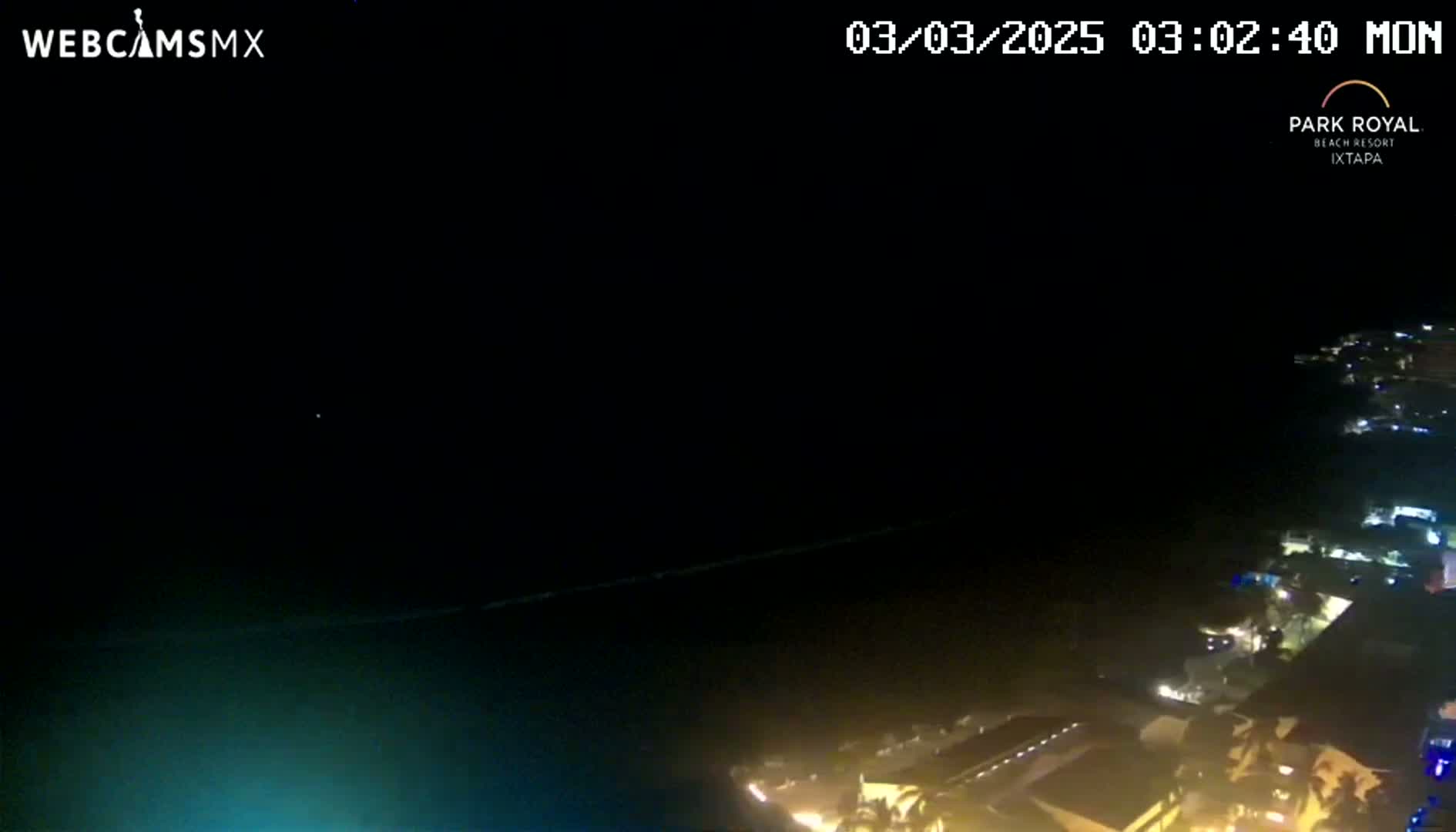 A nighttime, high-angle view shows a dark body of water with a coastline of illuminated buildings and vegetation under a clear, dark sky.