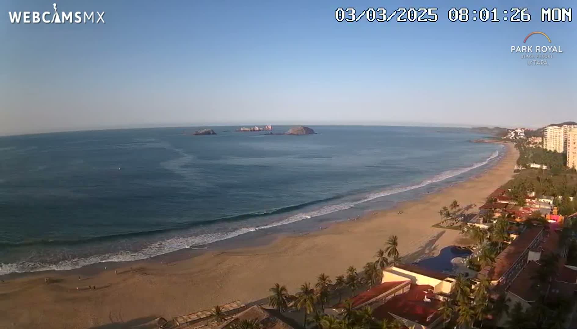 A beach with calm waves under a clear blue sky is viewed from above, showing a resort and several small islands in the distance.