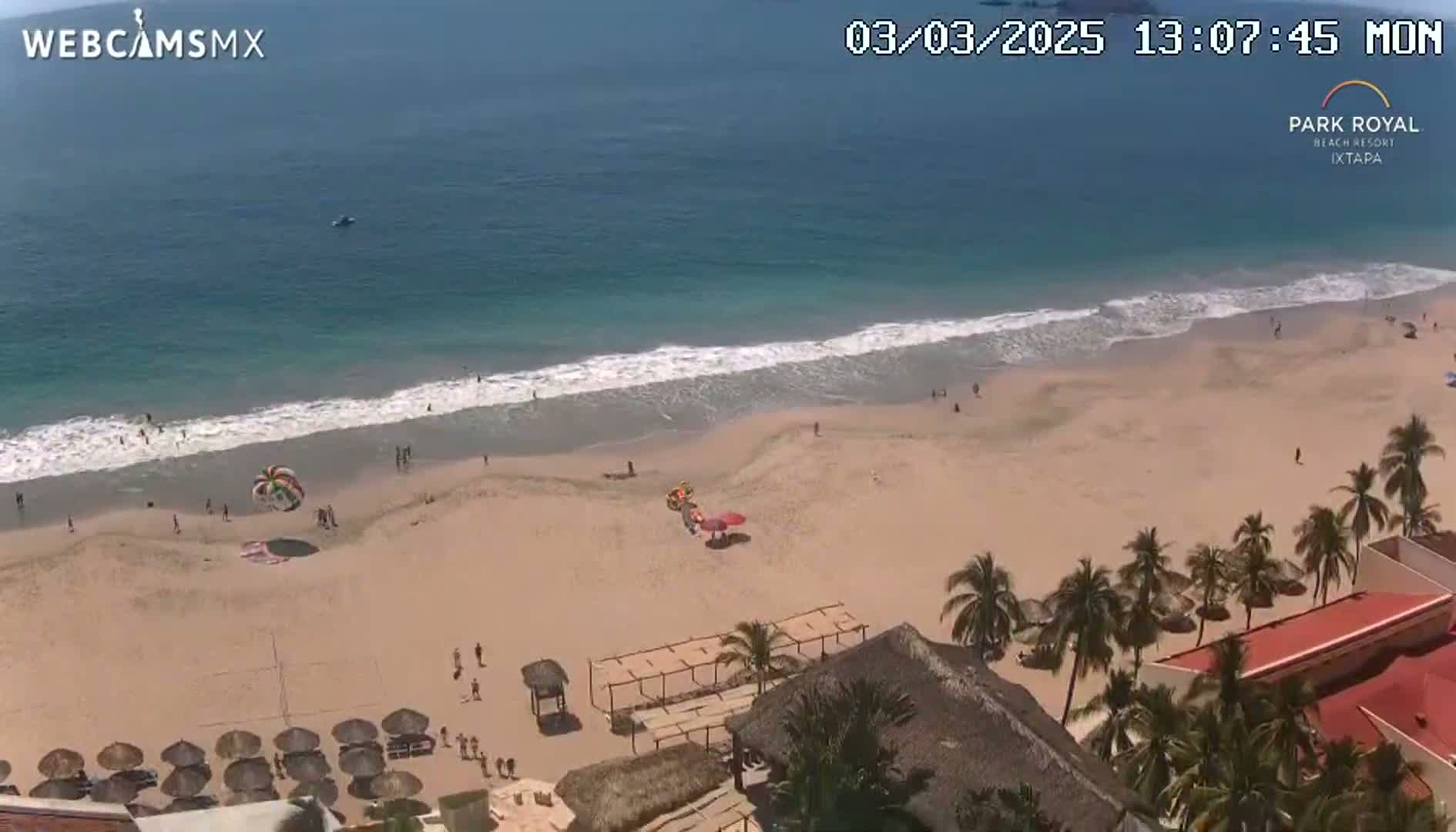 An aerial view shows a sandy beach with calm waves and a few people, parasailing equipment, and beach umbrellas under a sunny sky, next to a resort with palm trees.