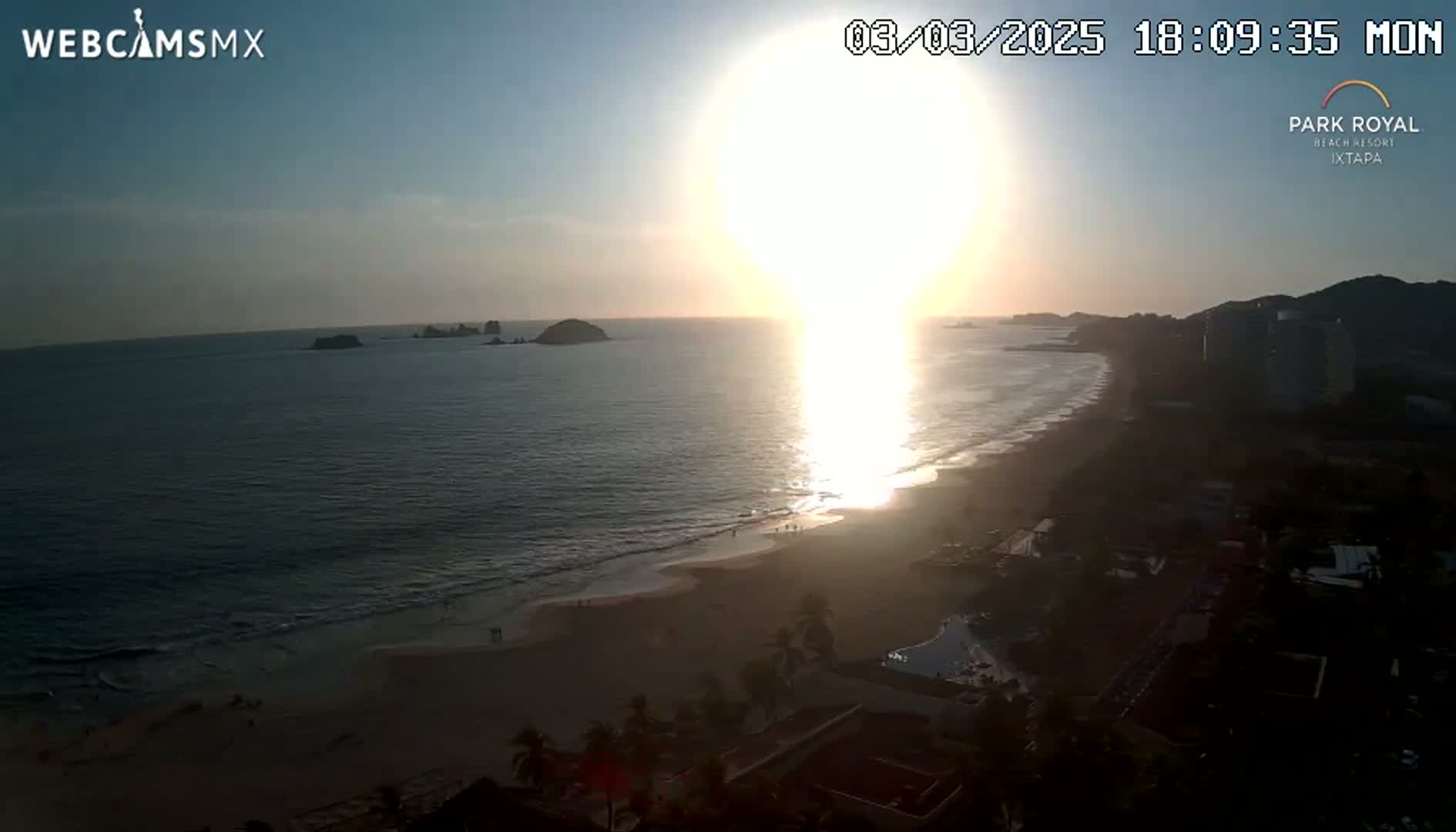 A bright sunset over a sandy beach with calm ocean waves and distant rocky islands, viewed from an elevated position overlooking coastal buildings.