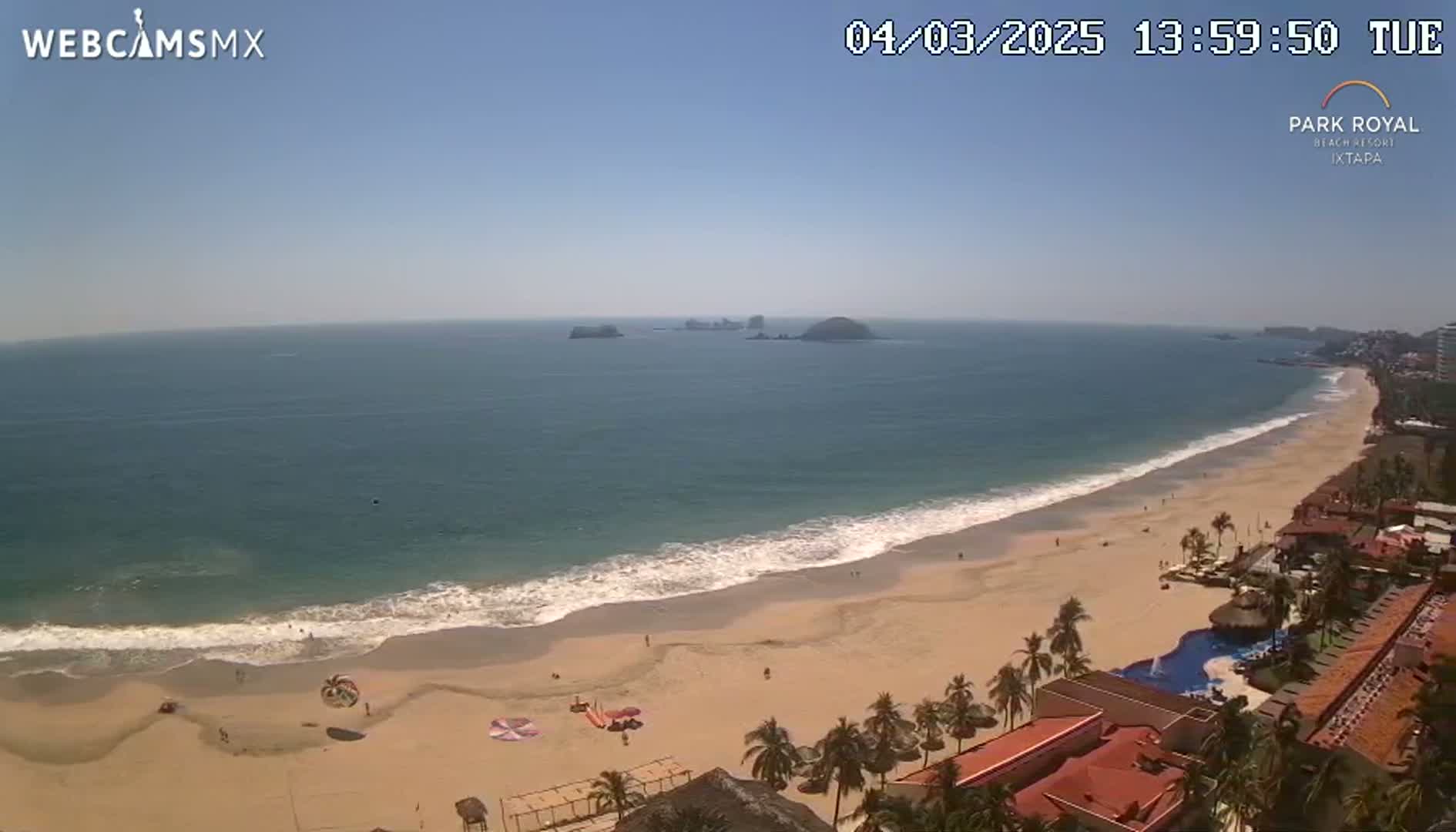 An aerial view shows a long, sandy beach with gentle waves lapping the shore under a clear, sunny sky, and a resort complex along the beach.