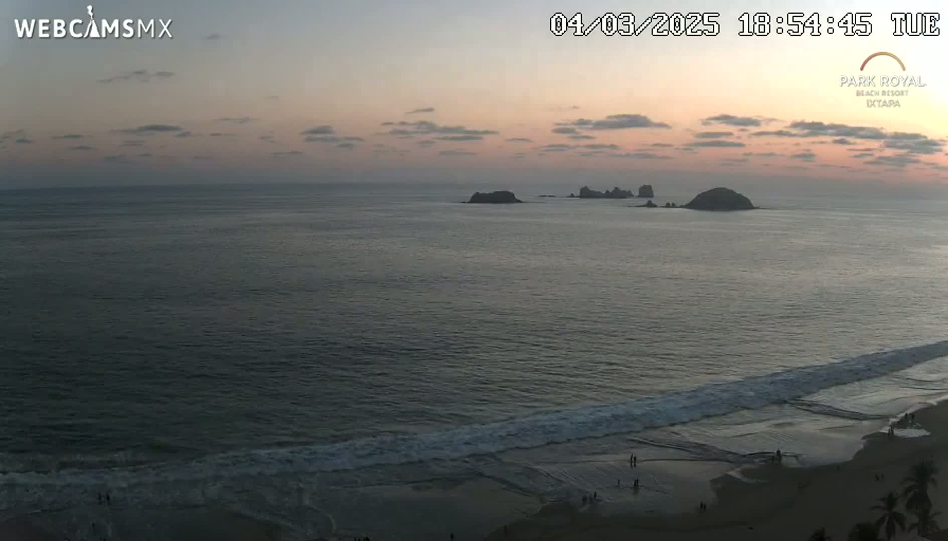 A tranquil ocean scene at sunset shows a sandy beach with small waves rolling in, several dark-colored islands in the distance, and a few people visible on the beach.