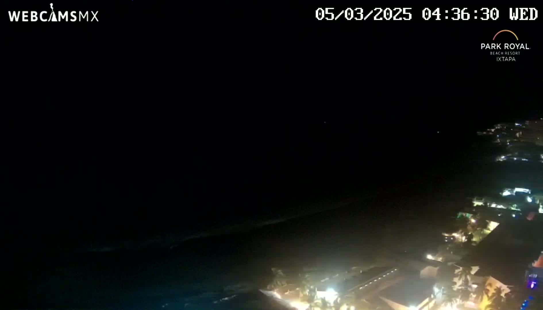 A nighttime coastal view shows a dark ocean with a dark sky, and a dimly lit shoreline with buildings and lights.