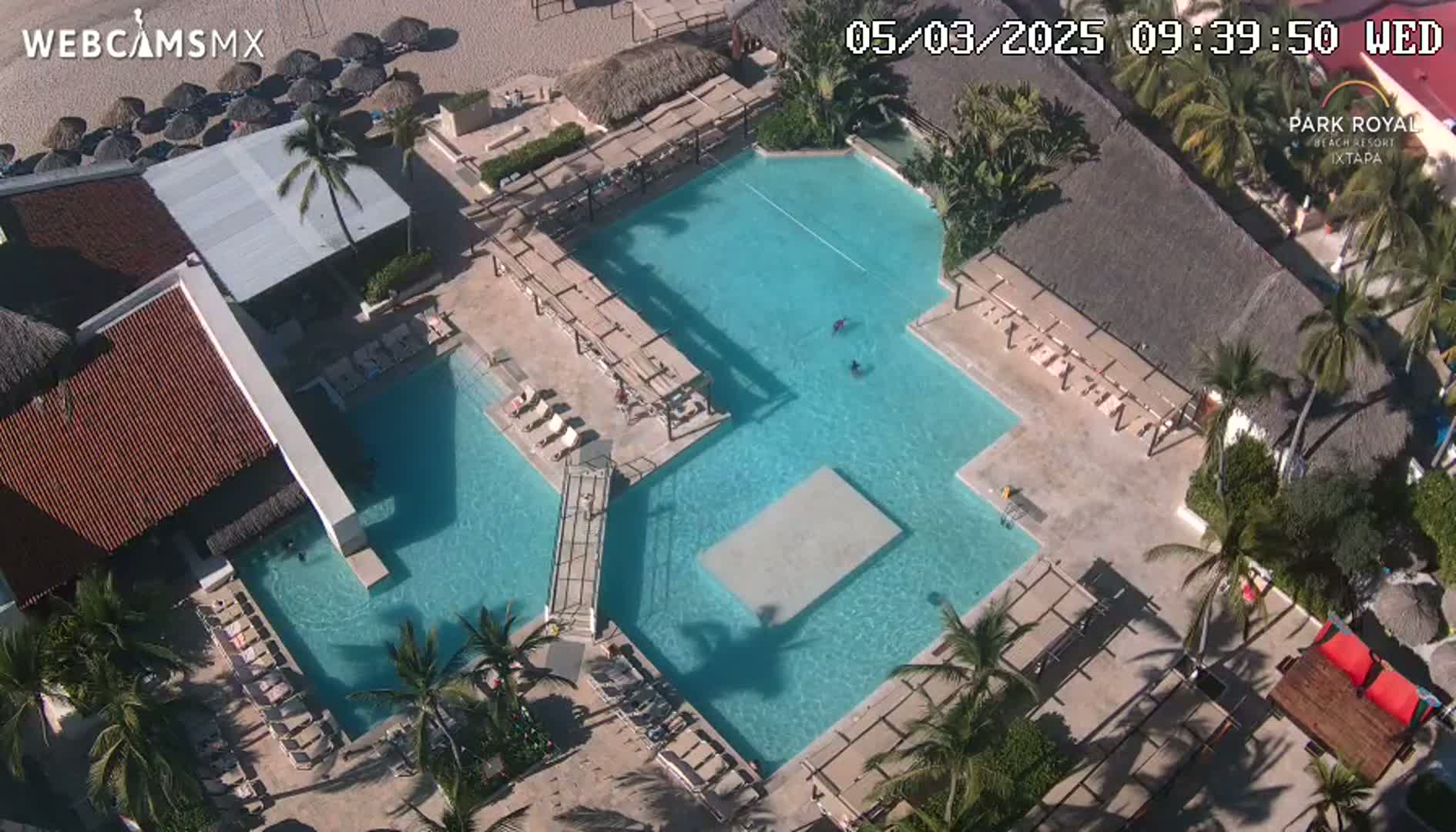 An aerial view shows a resort pool complex with several interconnected pools, lounge chairs, palm trees, and buildings under sunny conditions.