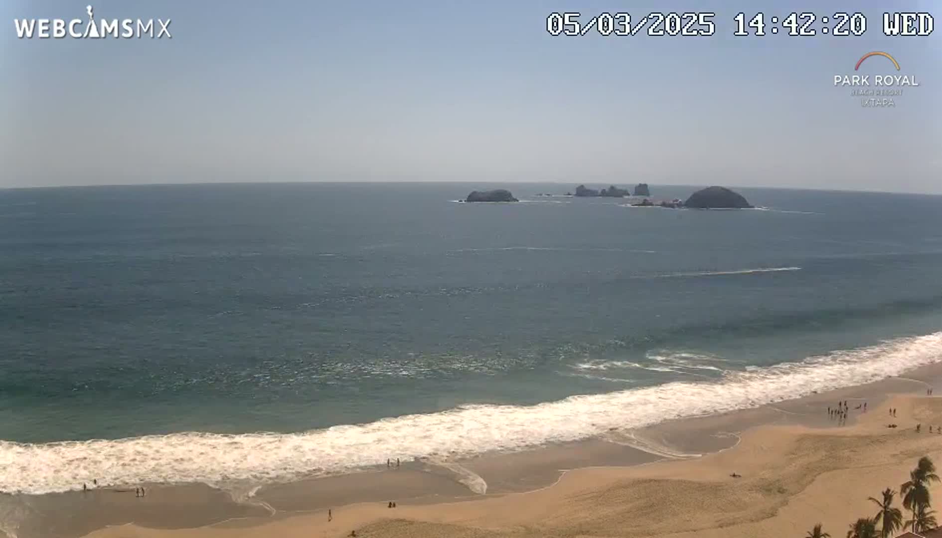 A mostly sunny day shows a sandy beach with small waves lapping the shore, and several small islands visible in the distance.