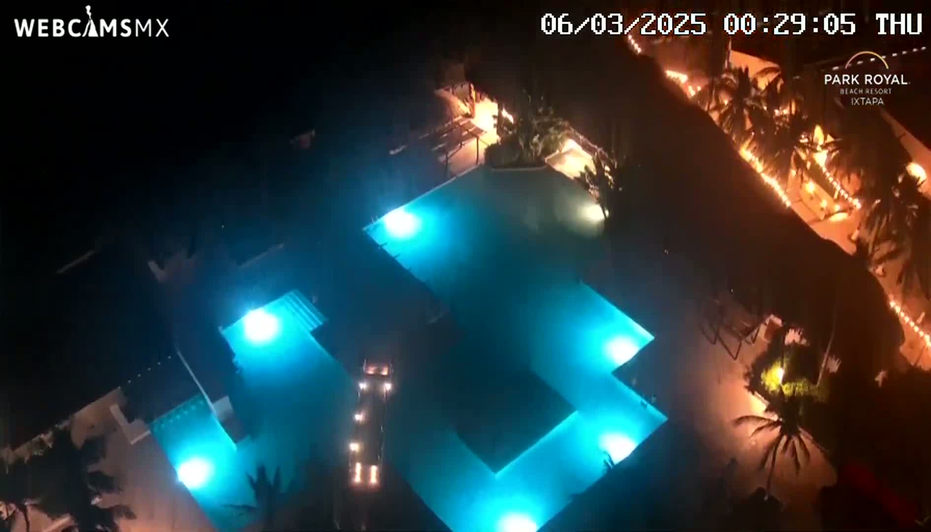 An overhead nighttime view shows a brightly lit, turquoise-blue pool complex surrounded by palm trees and buildings.