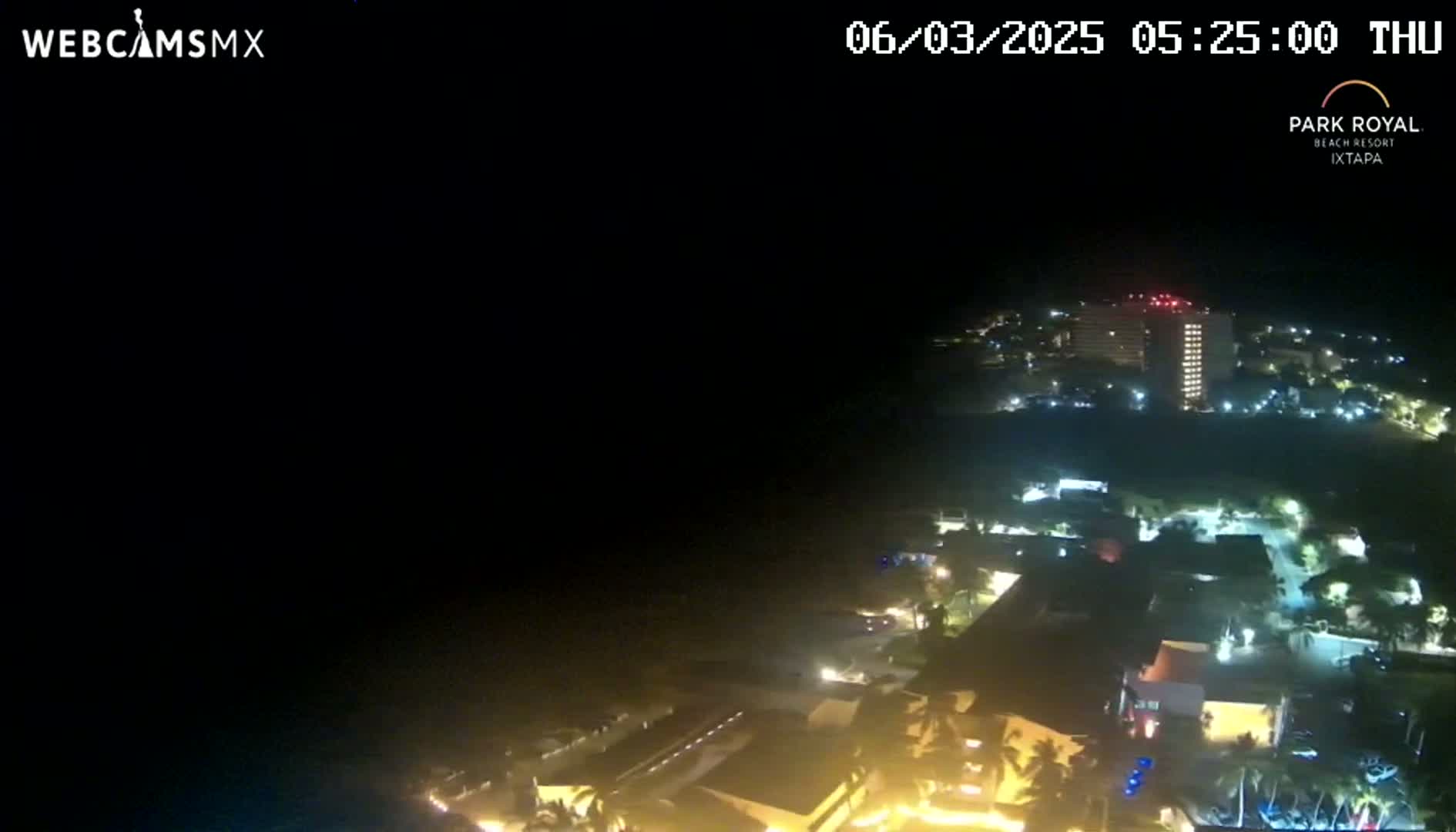 A nighttime aerial view shows a coastal town with illuminated buildings and streets, under a clear, dark sky.