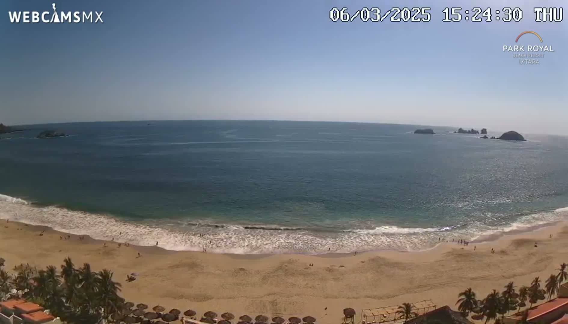 A sandy beach with gentle waves laps the shore under a clear blue sky, with a few people visible on the beach and distant islands in the ocean.
