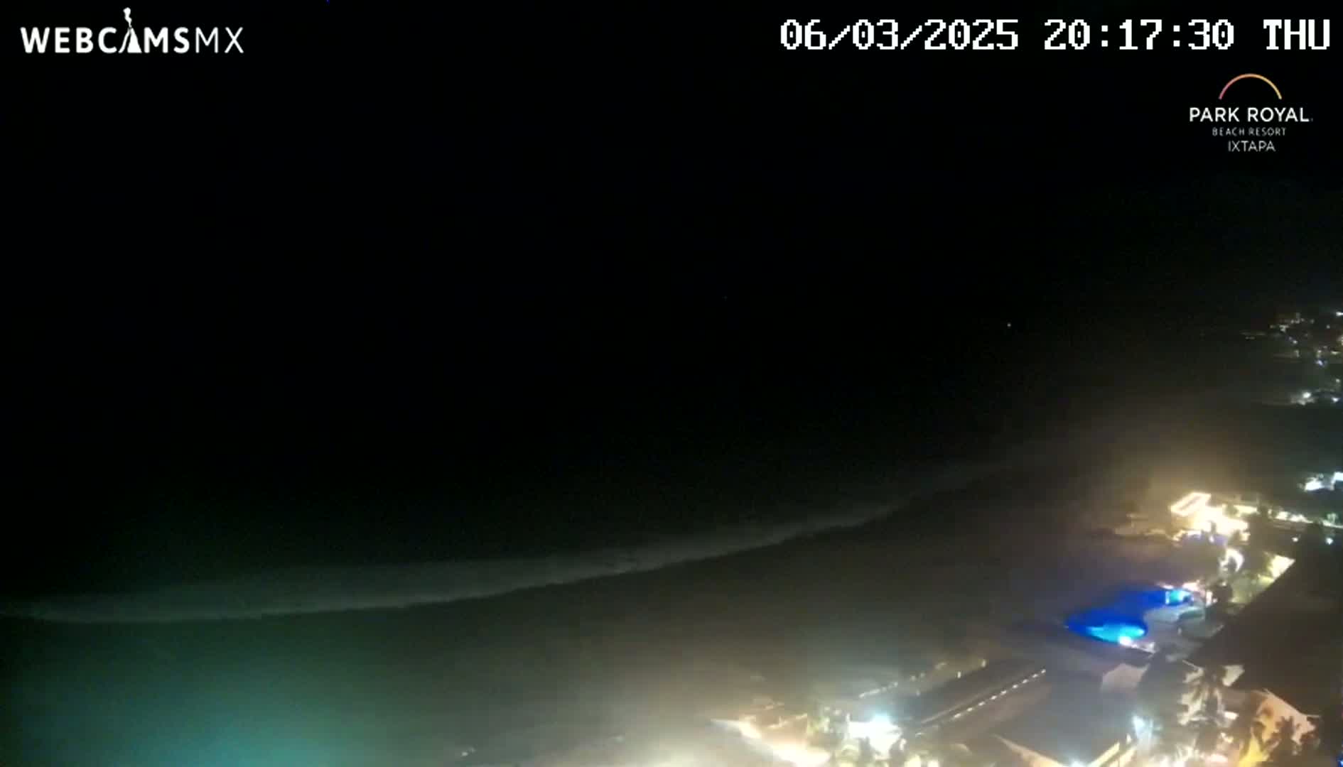 A nighttime, high-angle view shows dark ocean waves lapping a shore illuminated by the lights of a coastal resort.