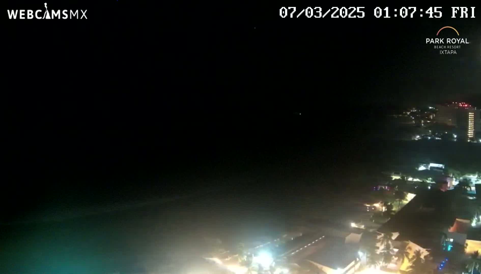 A nighttime, overhead view shows a dark ocean, dimly lit beach, and buildings with lights illuminating the scene.