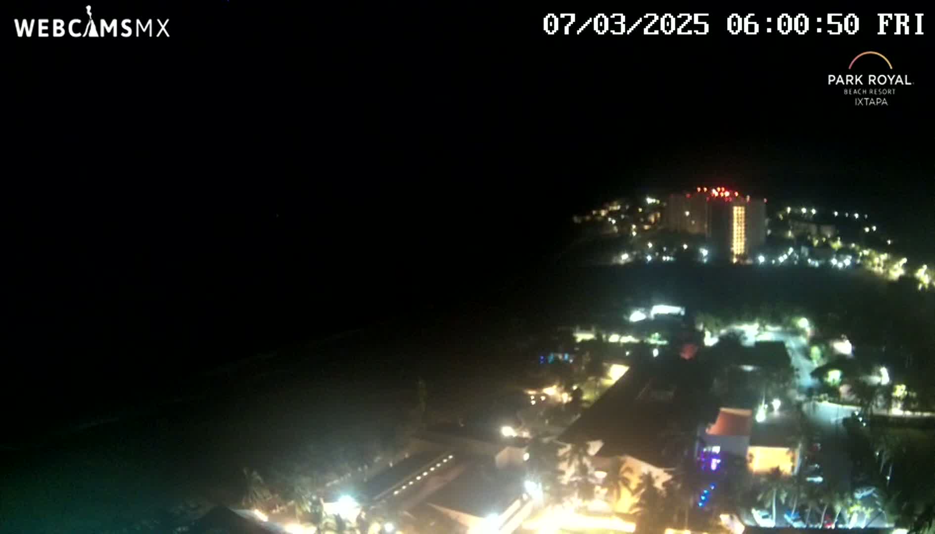 A nighttime, aerial view shows a coastal resort town with many lights illuminating buildings and streets, and a dark ocean in the foreground.