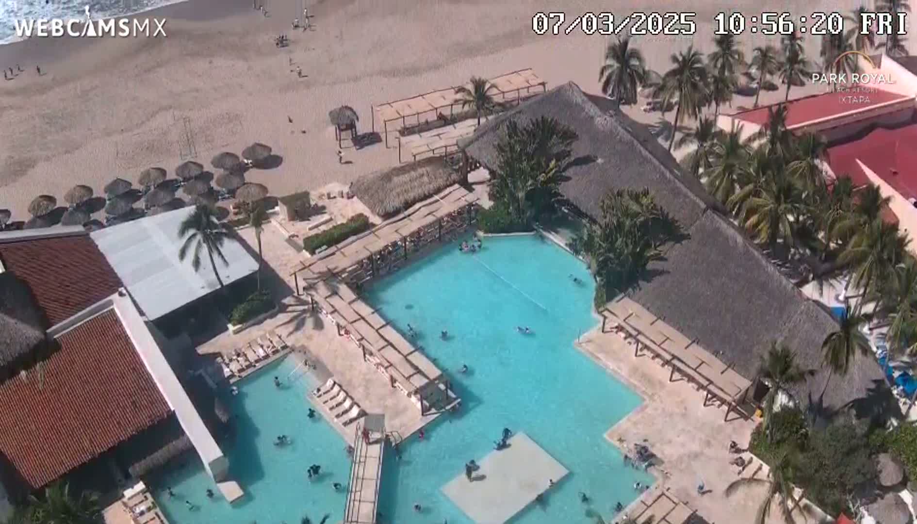 An aerial view shows a resort with a large, multi-level pool complex next to a sandy beach on a sunny day.