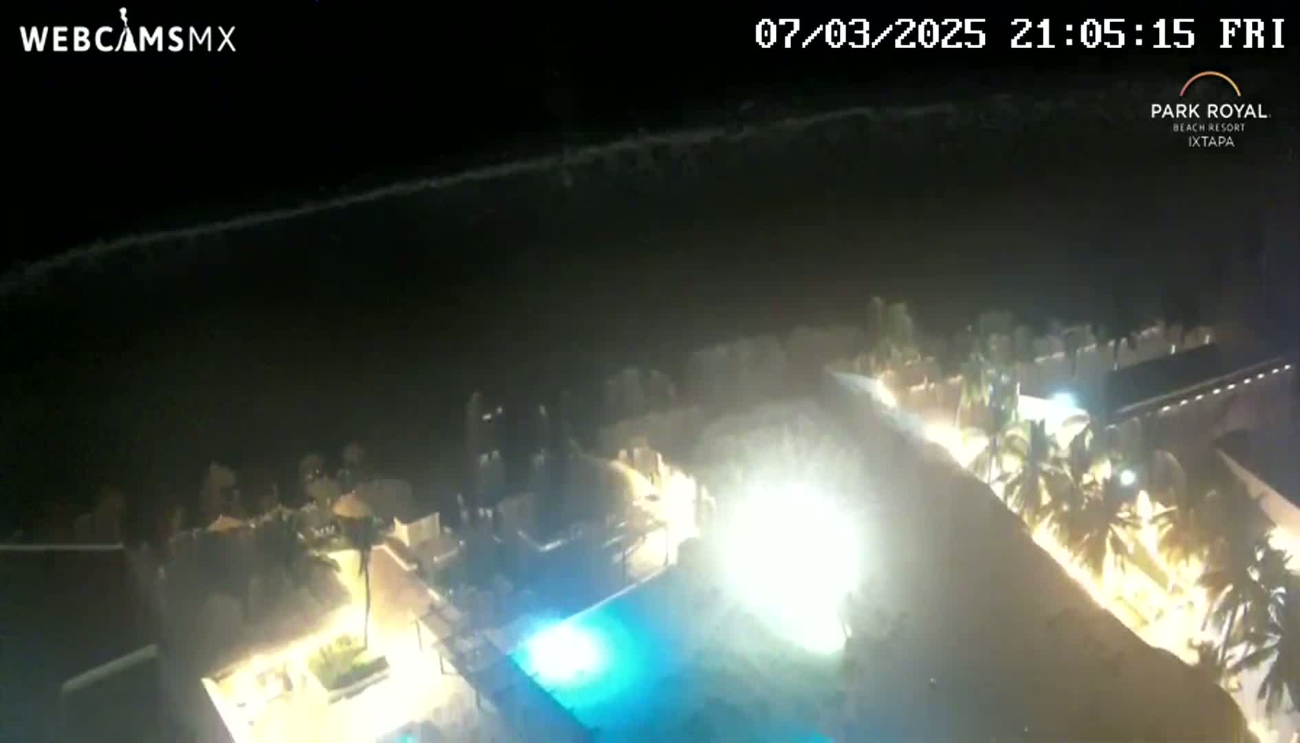 A nighttime aerial view shows a resort complex with a lit pool and palm trees near a dark ocean with waves under a clear sky.