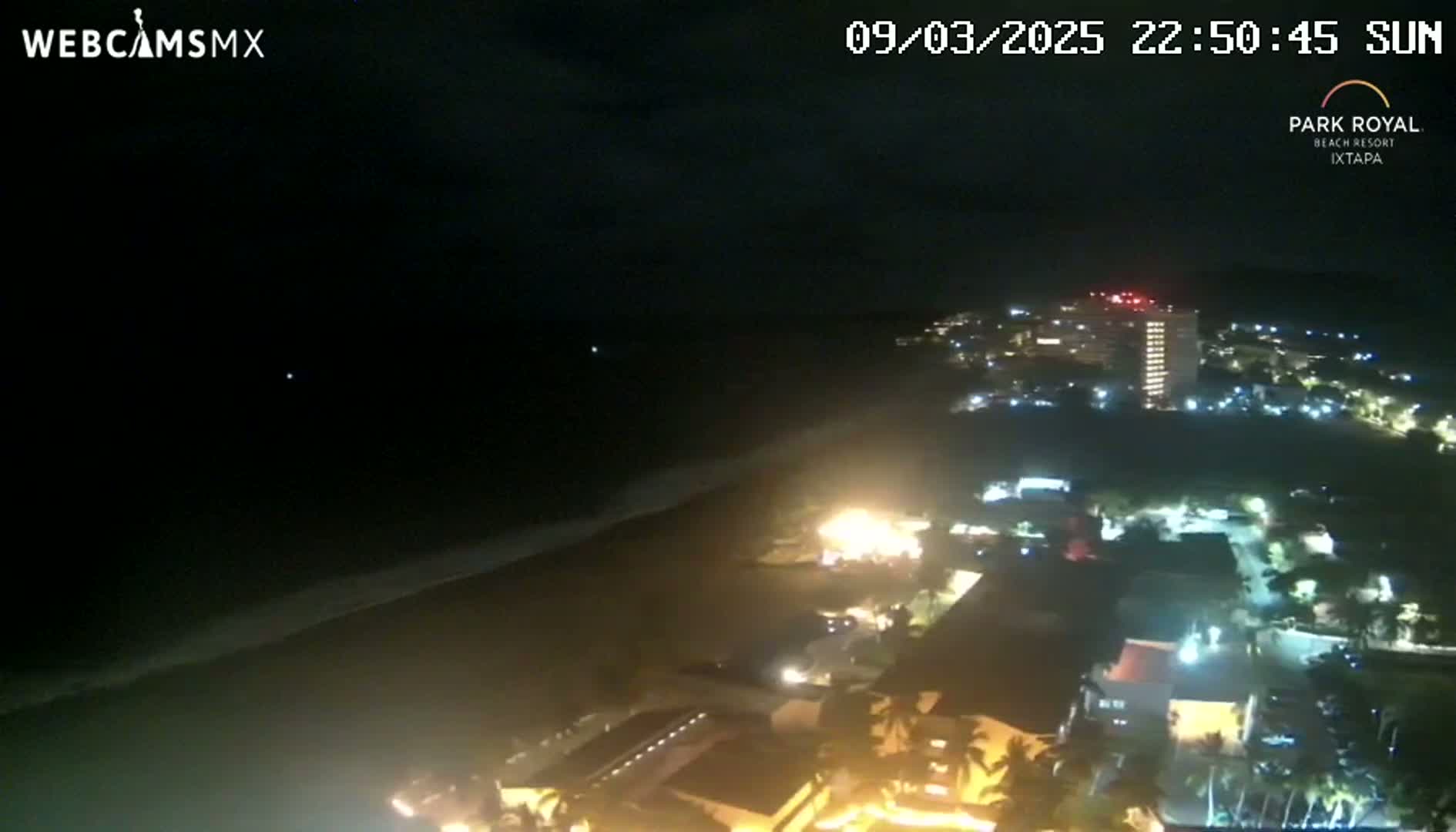 A nighttime aerial view shows a dark ocean with waves lapping a beach next to a brightly lit coastal town under a clear sky.