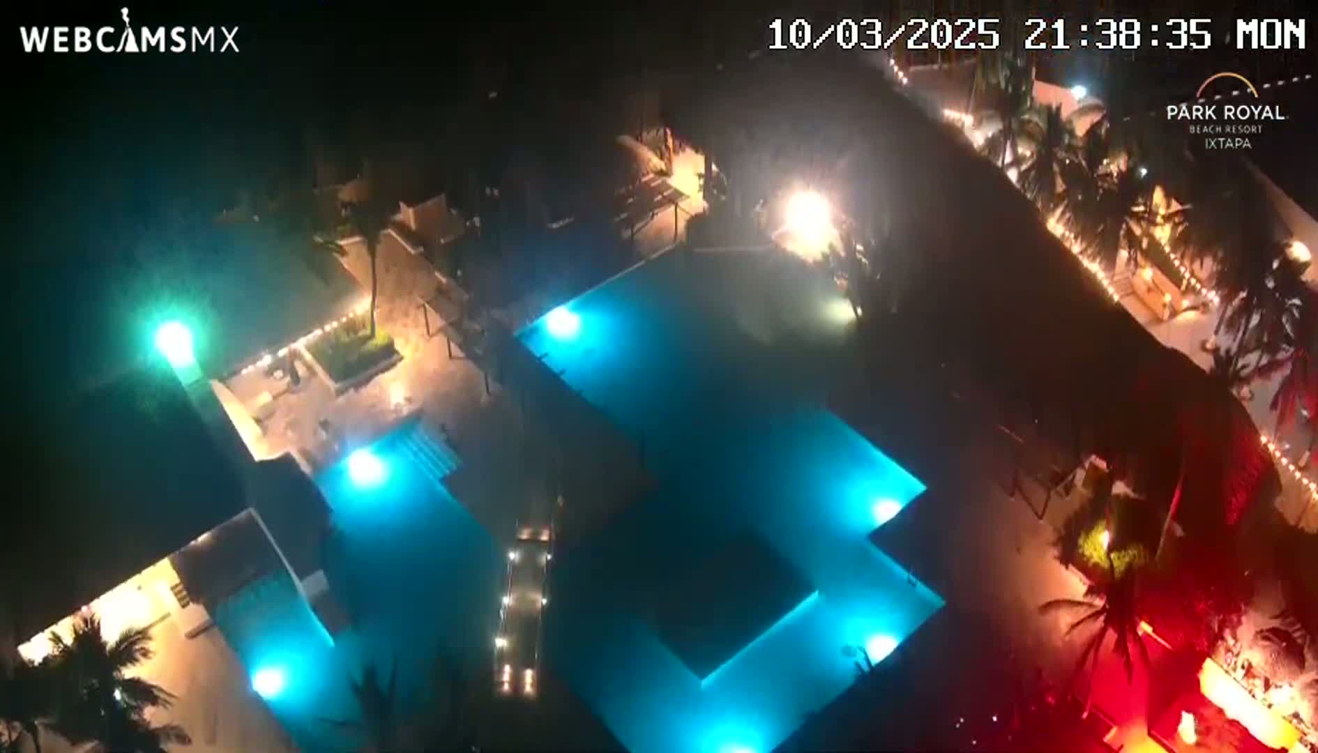 An aerial nighttime view shows two illuminated swimming pools at a resort, surrounded by palm trees and buildings.