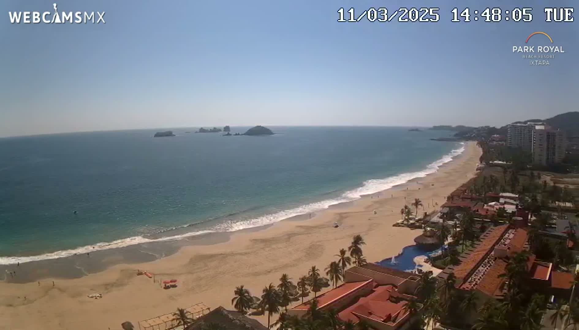 A sunny day at a beach with a long sandy shore, gentle waves, and a resort complex visible in the foreground.