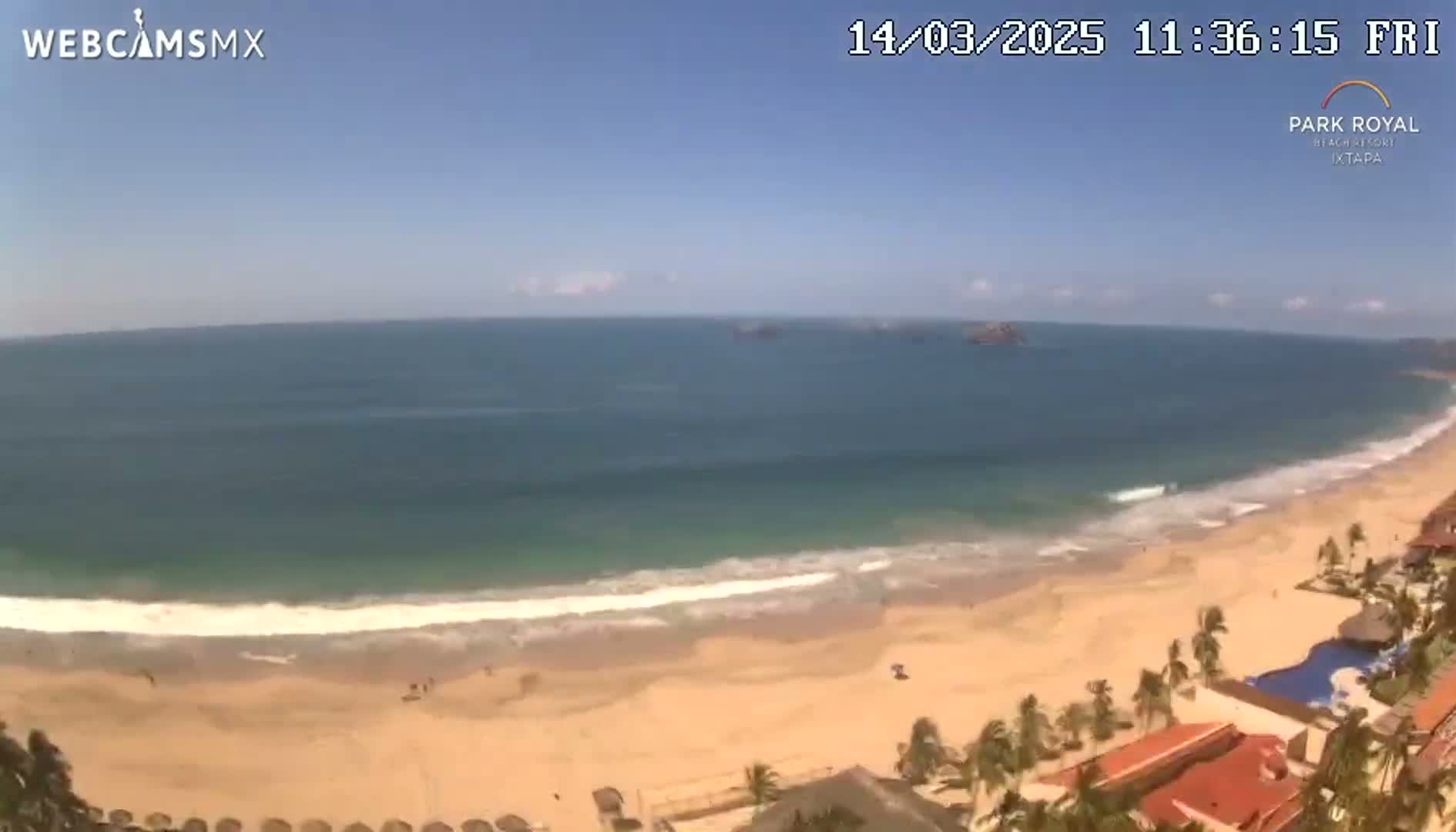 An expansive sandy beach meets a calm, blue ocean under a clear, sunny sky, with a resort partially visible in the foreground.