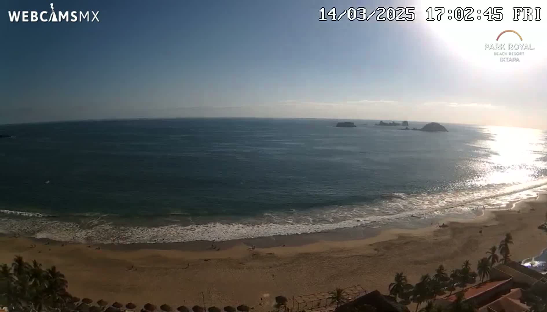 A sunny, high-angle view shows a sandy beach with gentle waves lapping the shore, and a calm ocean extending to the horizon with several small islands visible in the distance.