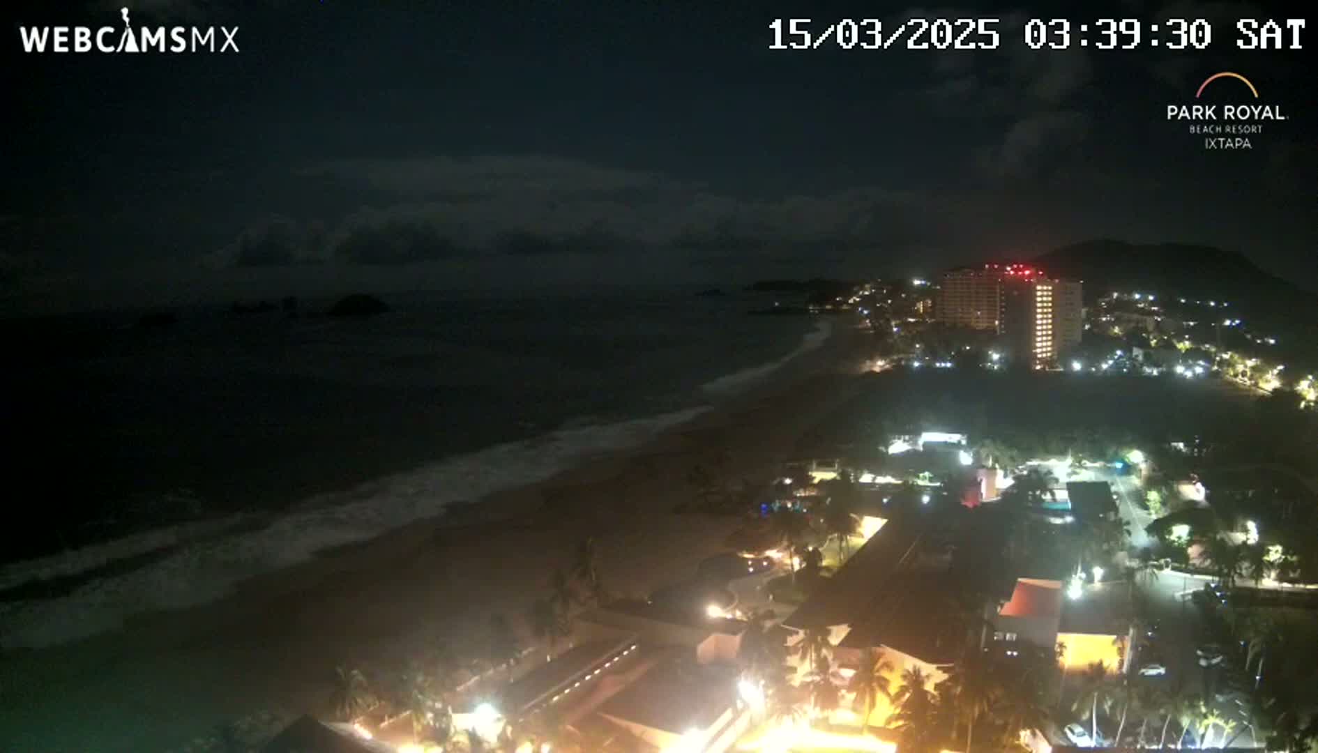A nighttime coastal scene shows a dark ocean with waves lapping a sandy beach, next to a city with illuminated buildings under a partly cloudy sky.