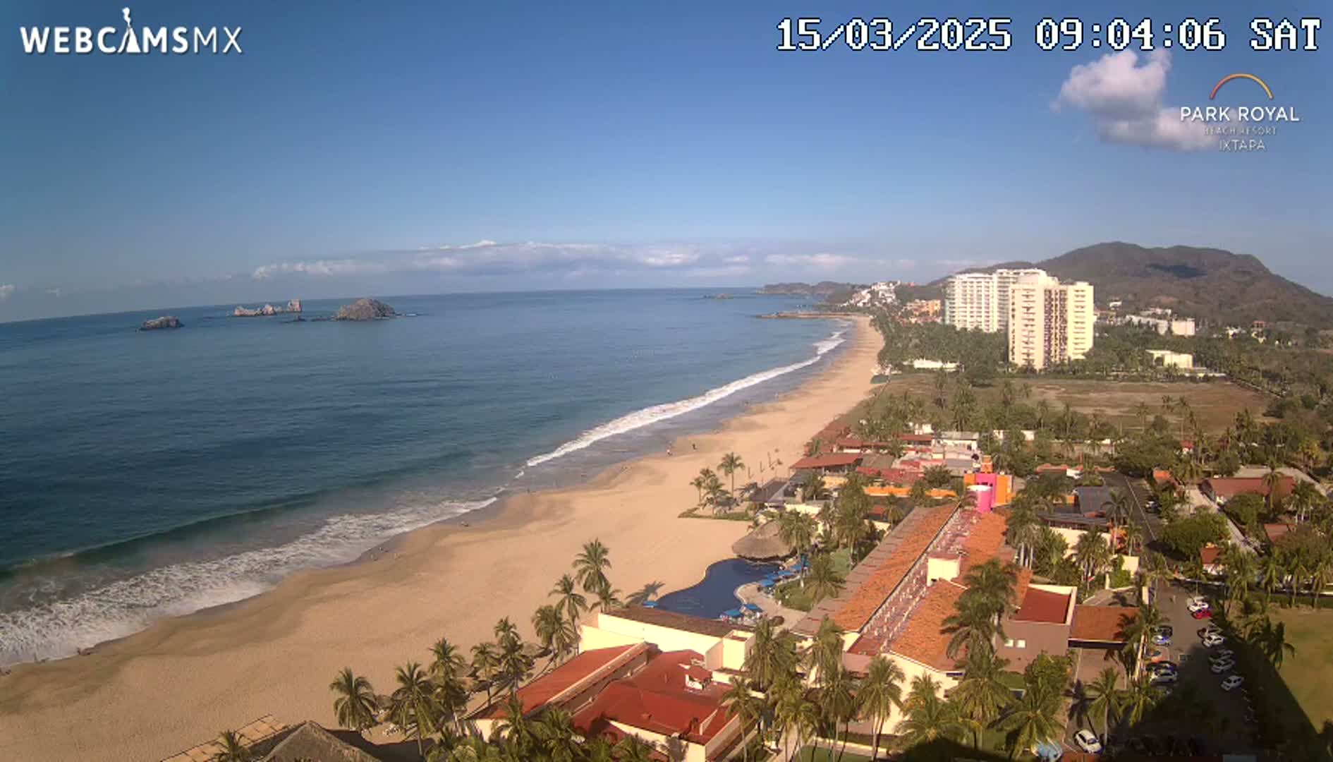 An aerial view shows a long sandy beach with gentle waves lapping the shore, under a clear blue sky, with resort buildings and palm trees visible along the beachfront and a mountainous backdrop.