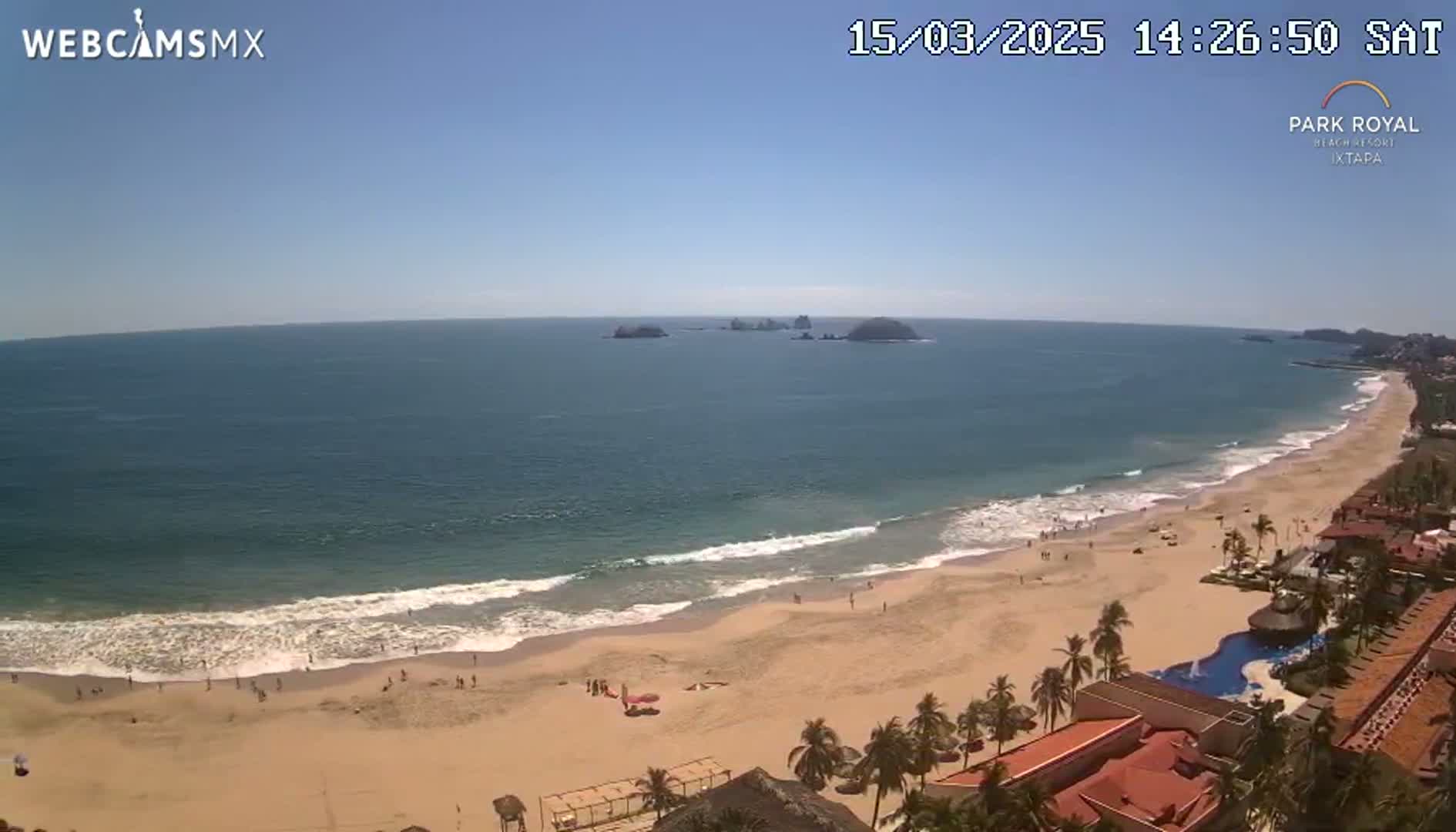 An expansive sandy beach with gentle waves under a clear blue sky, overlooks a calm ocean with distant islands and a resort in the foreground.