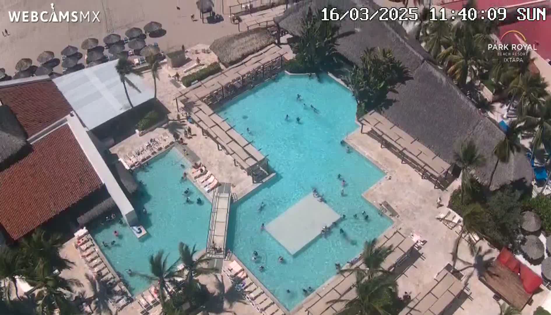 An aerial view shows a resort pool complex with numerous people swimming and lounging on sunny day.