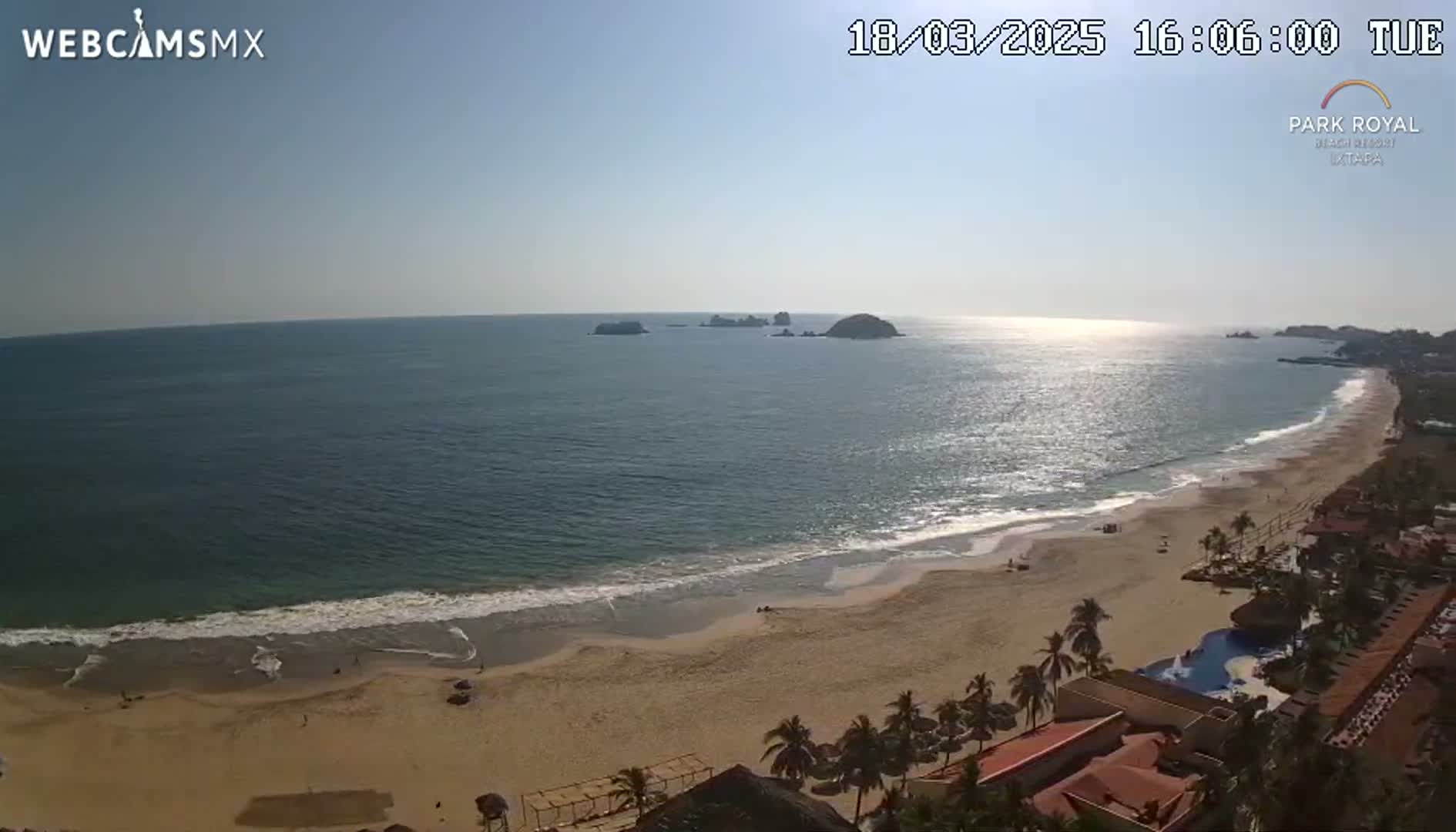 An expansive sandy beach meets a calm ocean under a clear, sunny sky, with distant islands visible and resort buildings in the foreground.