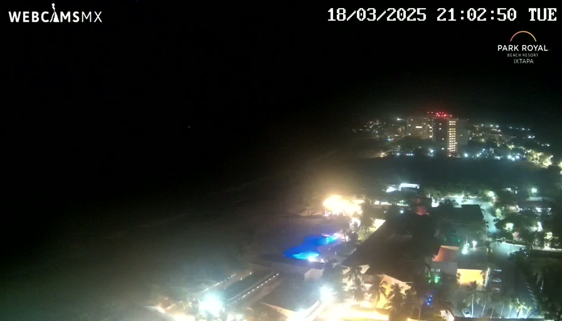 A nighttime, aerial view shows a coastal resort town with its lights illuminating the beach and buildings under a clear, dark sky.