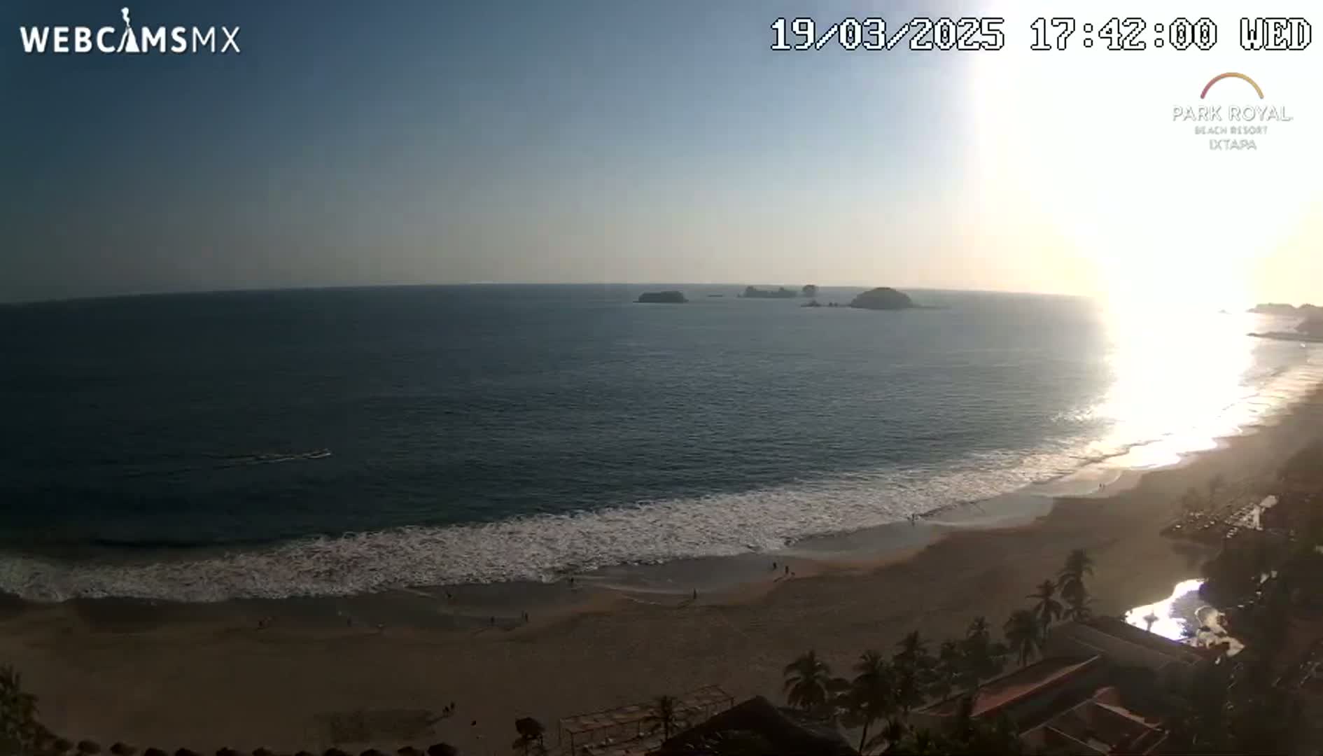 A beach with calm waves and a sunset over the ocean, with islands in the distance.