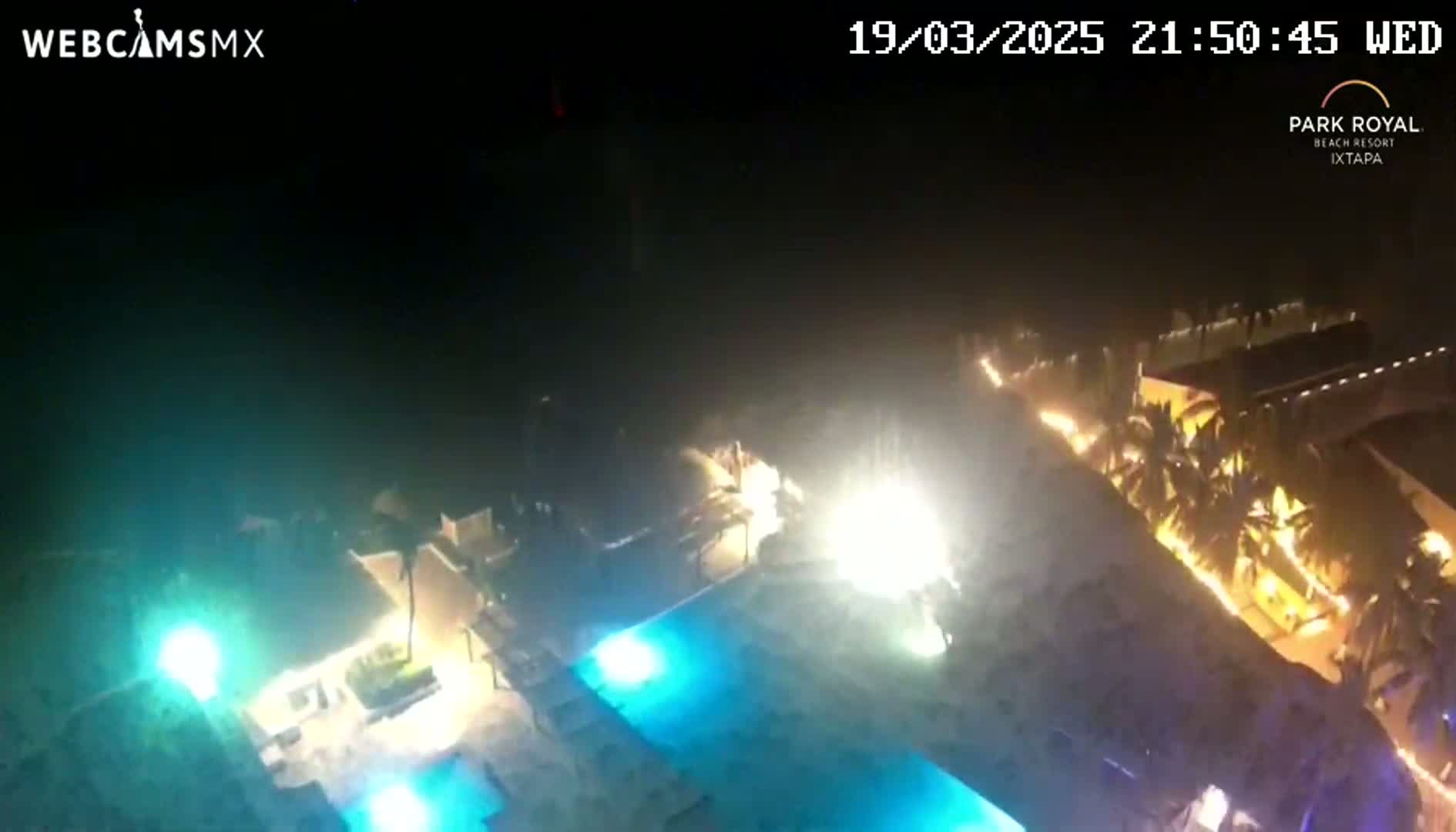 A nighttime aerial view shows a resort with illuminated pools and palm trees under a dark, clear sky.