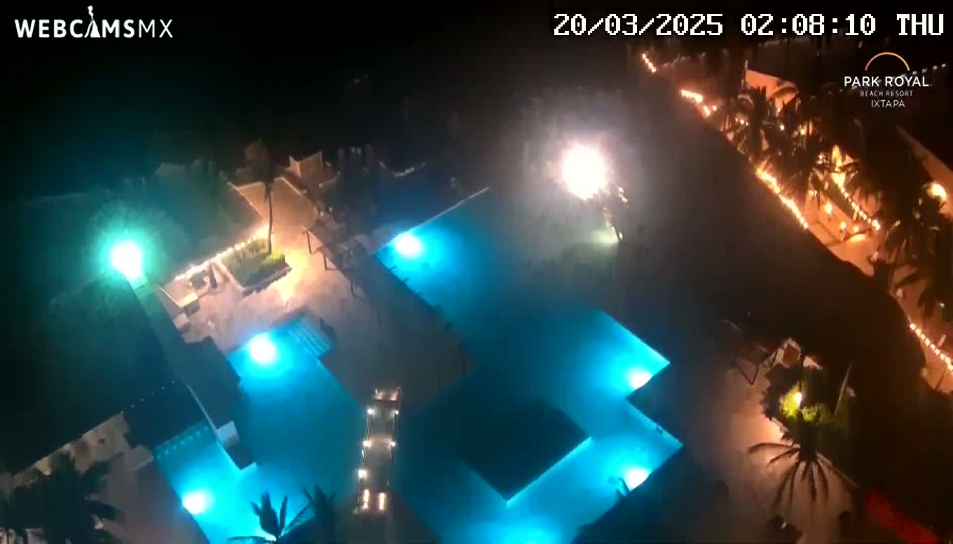 An aerial nighttime view shows a resort with illuminated pools and surrounding buildings, under a clear sky.