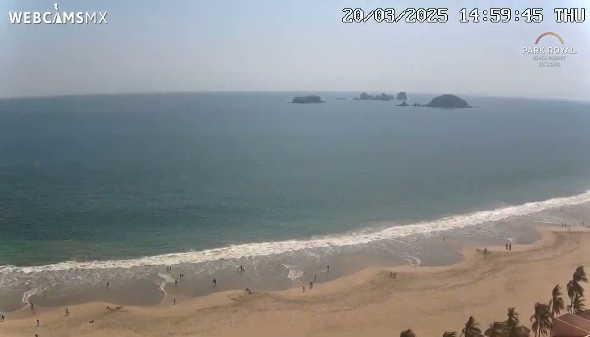 A sandy beach with a few people on it slopes down to a calm ocean with several small islands visible in the distance under a hazy sky.