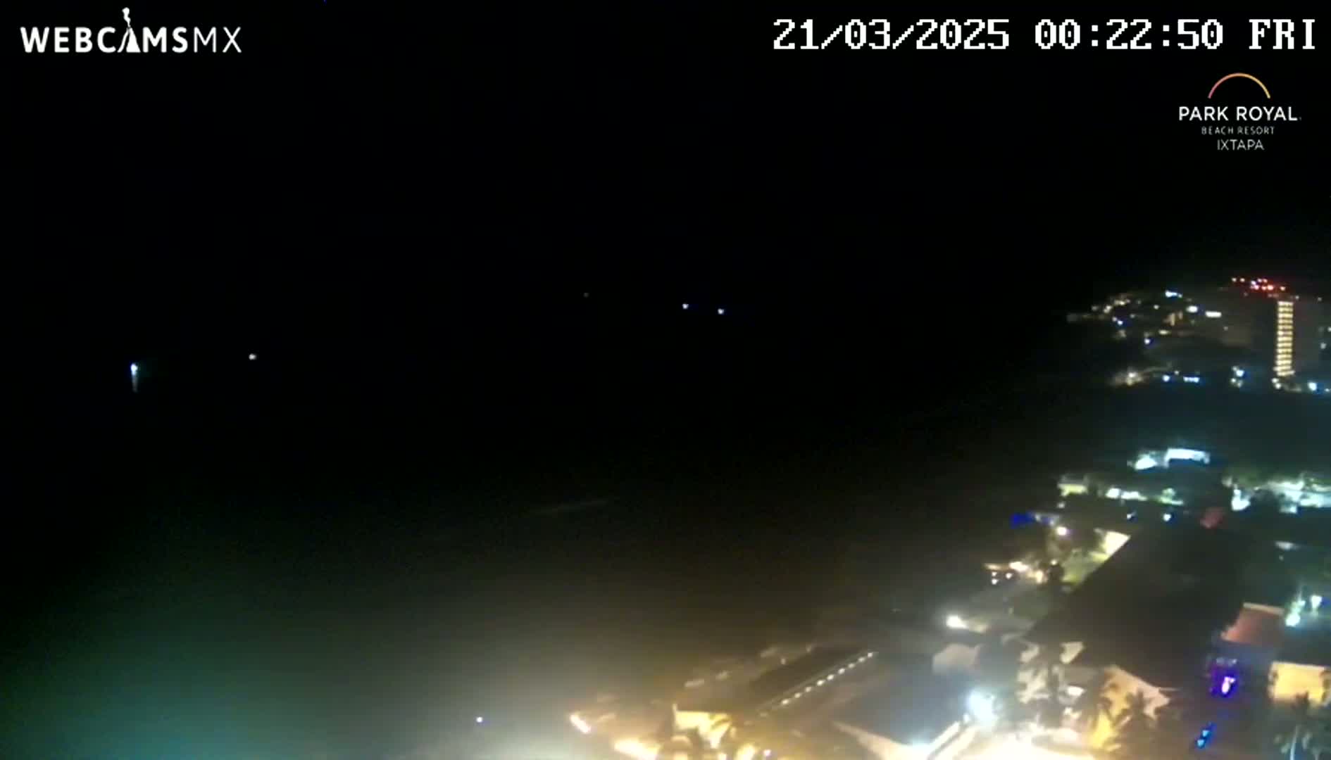A nighttime, high-angle view shows a dark ocean and a brightly lit coastal town with buildings and streetlights.