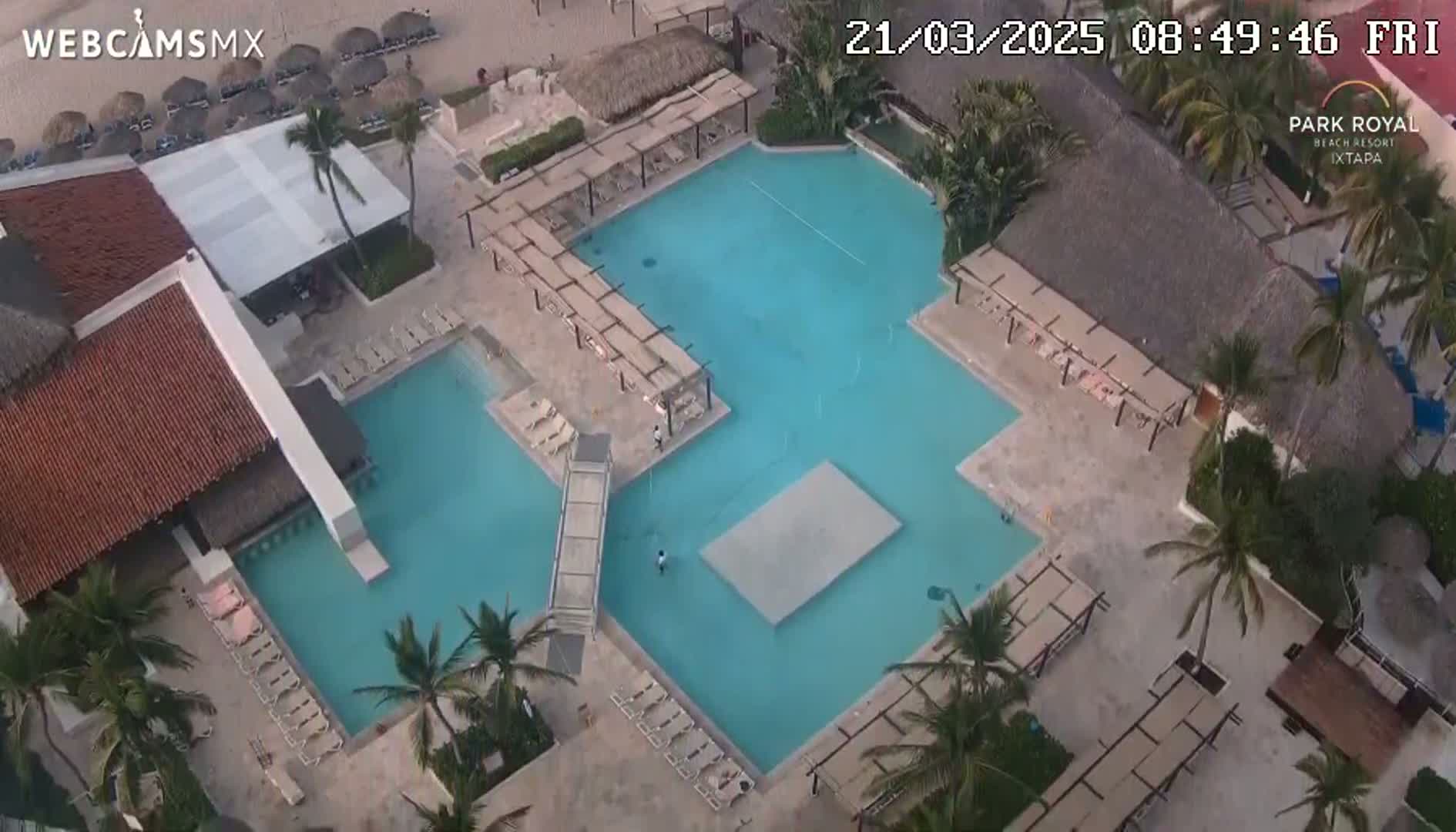 An aerial view shows a large, light-blue resort pool complex with surrounding lounge chairs and palm trees on a sunny day.