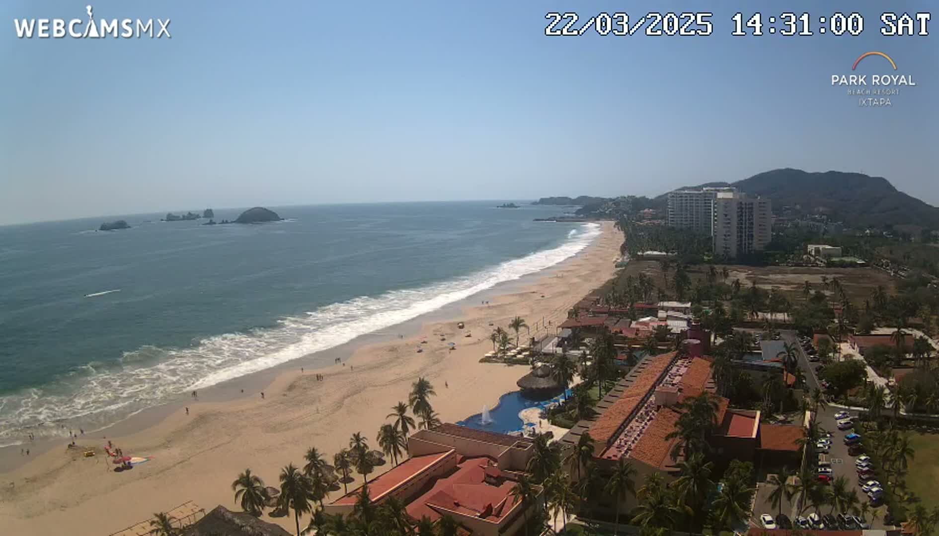 An aerial view shows a long sandy beach with gentle waves lapping the shore under a clear blue sky, beachfront hotels and resorts, and a mountain in the distance.