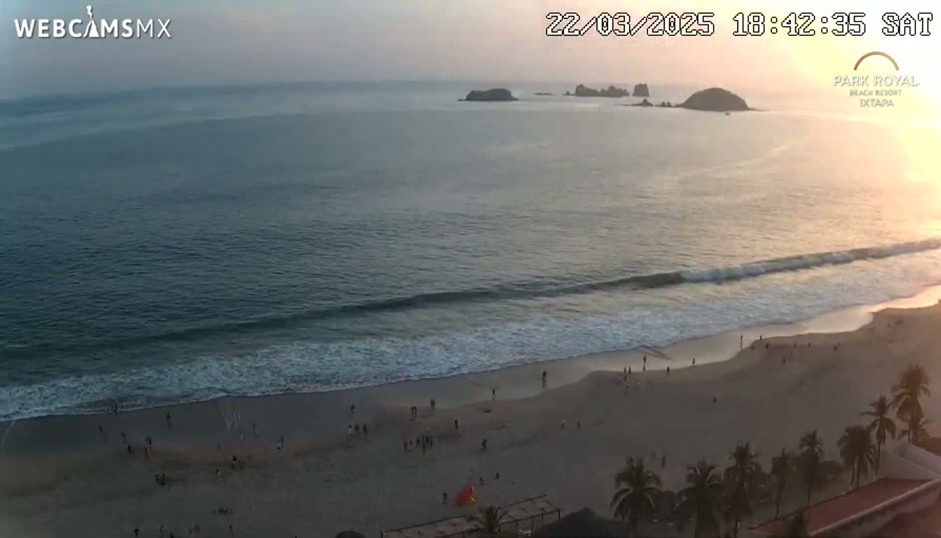 An aerial view shows a beach with people on it, gentle waves lapping the shore, and islands in the distance during what appears to be sunset.