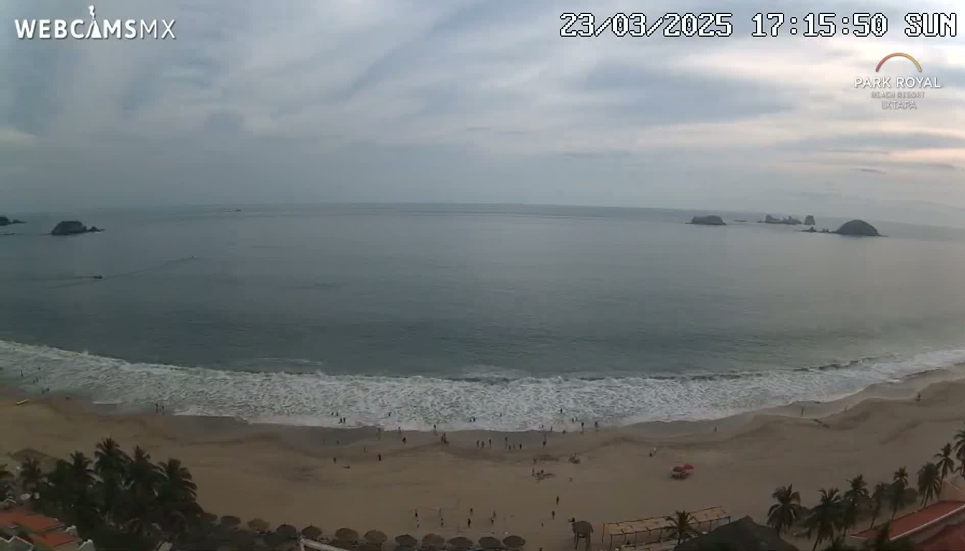 A sandy beach with gentle waves and several people on it, under an overcast sky, is visible from a high vantage point, with small islands visible in the distance.