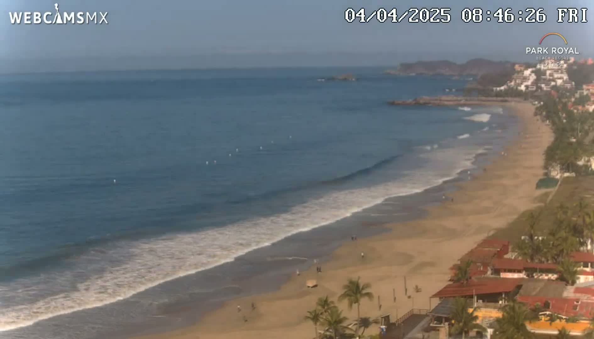 A long sandy beach with gentle waves and a few people, bordered by buildings and palm trees under a clear sky.