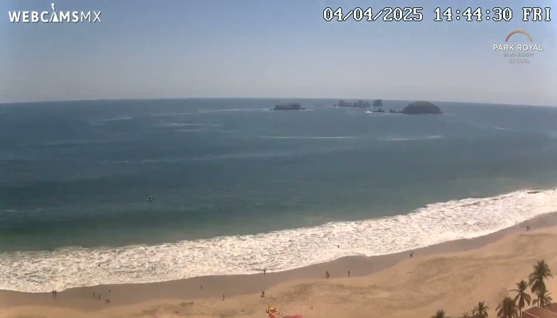 A sandy beach with gentle waves rolls onto the shore under a clear, sunny sky, with a few people visible on the beach and some islands in the distance.