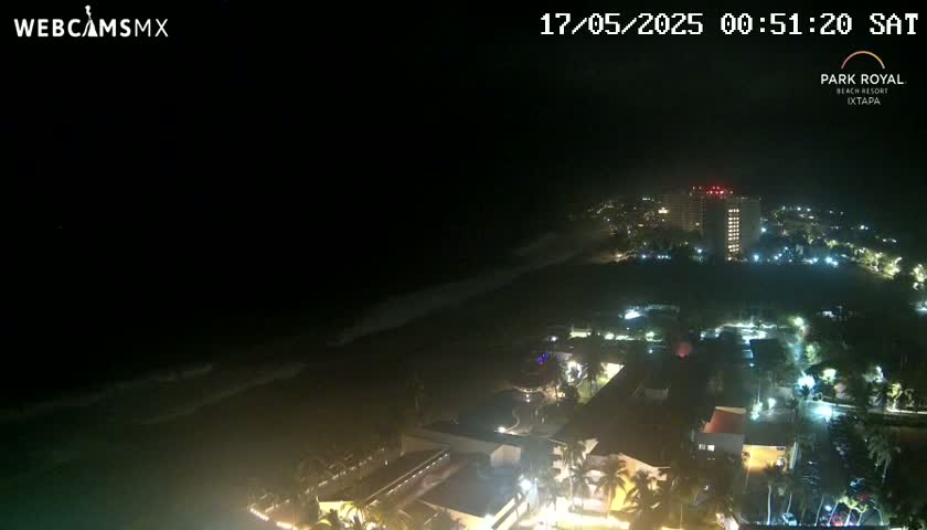 A nighttime coastal view shows a resort with illuminated buildings and palm trees, along with a dark ocean with gentle waves.